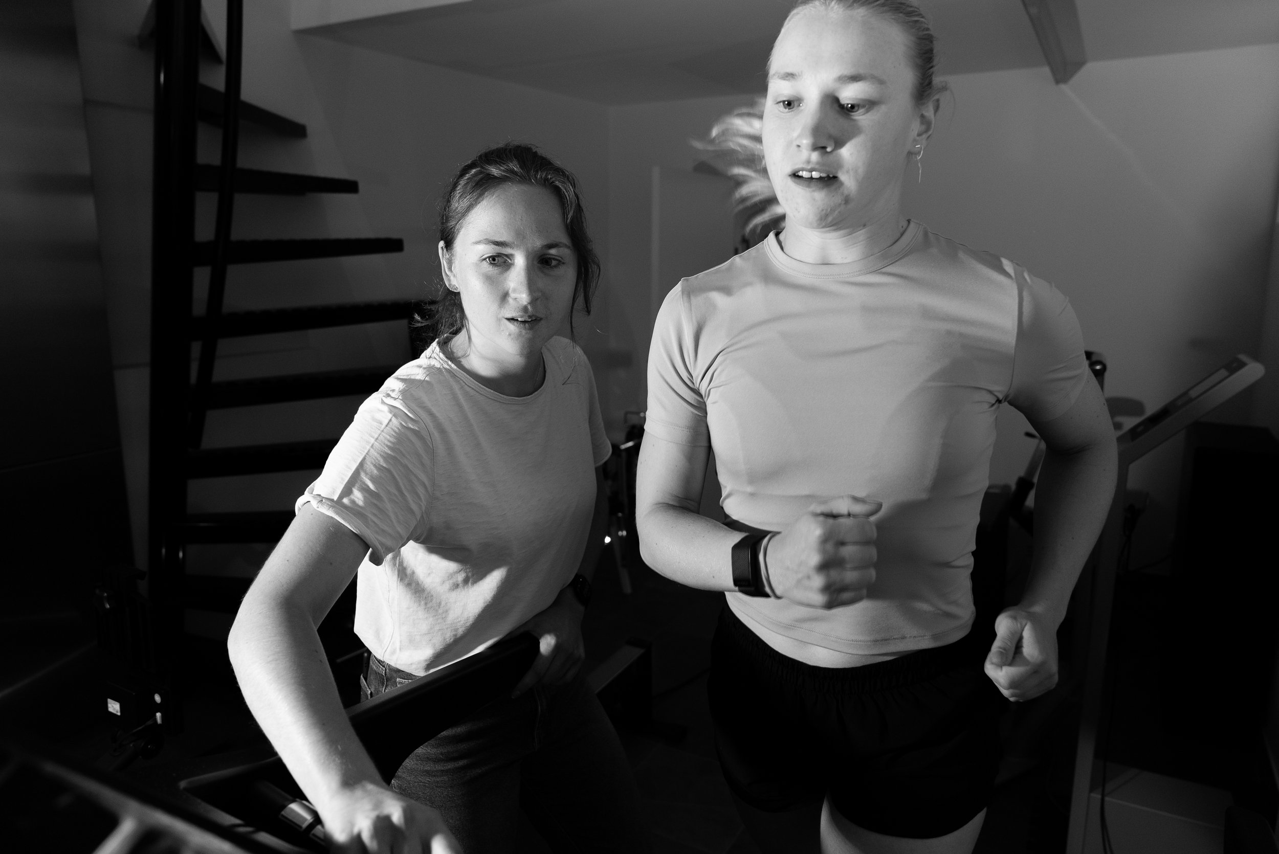 Twee vrouwen die hardlopen of joggen binnen, één kijkt naar de camera en de ander richt zich op het hardlopen, in een huis of sportschool, zwart-wil