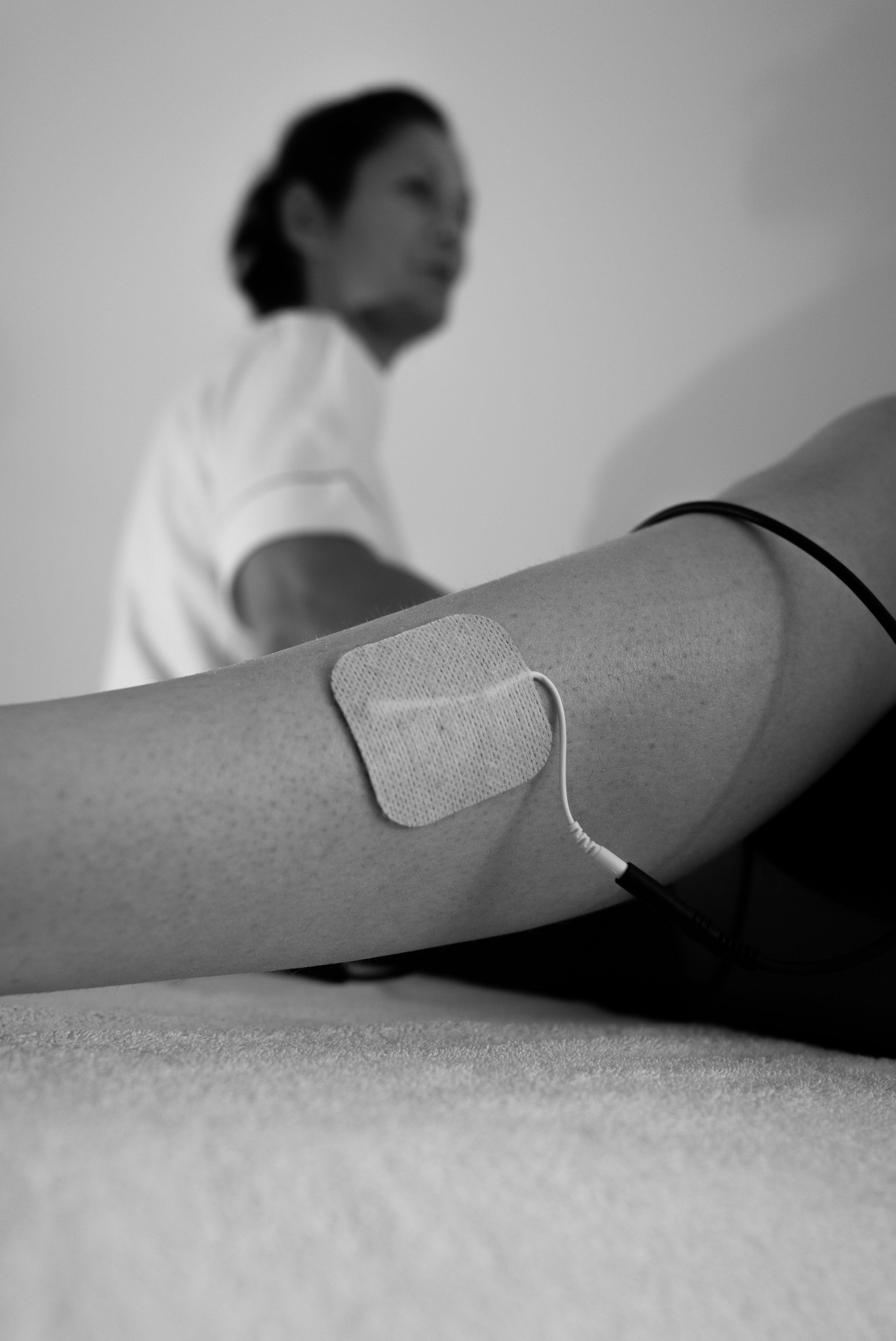 Close-up van een arm met een ECG-elektrode en kabel, met een persoon op de achtergrond die naar beneden kijkt.