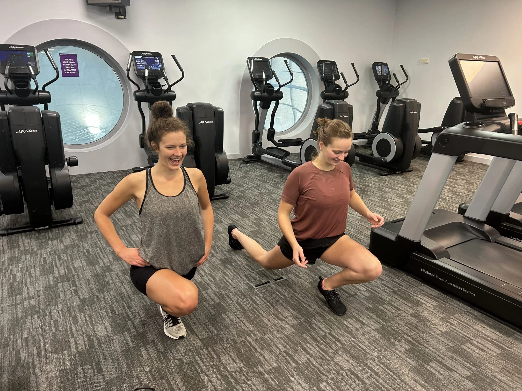 Two women in workout clothes are exercising in a gym with several elliptical machines in the background. One woman is standing and smiling while the other is kneeling and stretching her leg on a treadmill.