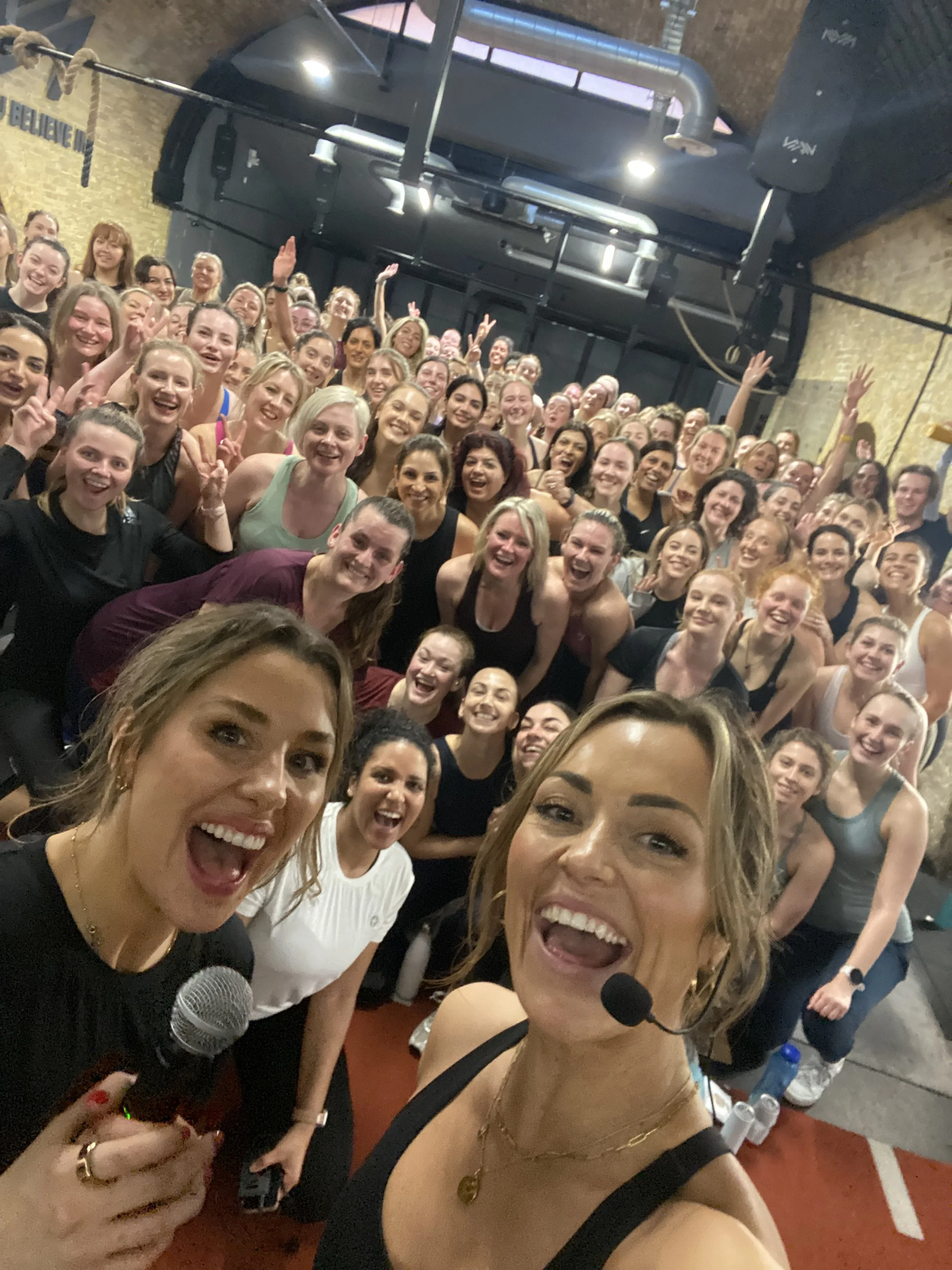 Two fitness instructors taking a selfie in front of a large group of women in workout attire, smiling and celebrating after a fitness class inside a gym.