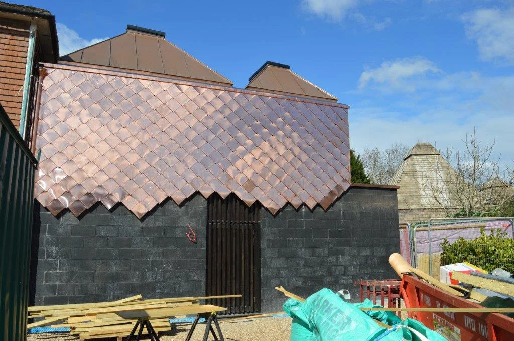 Building under construction with copper roof tiles in process of installation. Construction materials and tools are visible in the foreground.