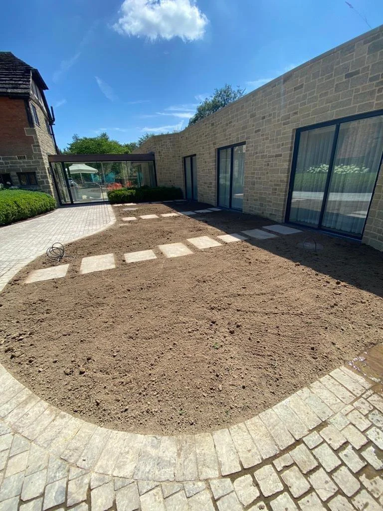 A backyard with a stone pathway and a partially landscaped area with dirt, next to a brick building with large glass doors.