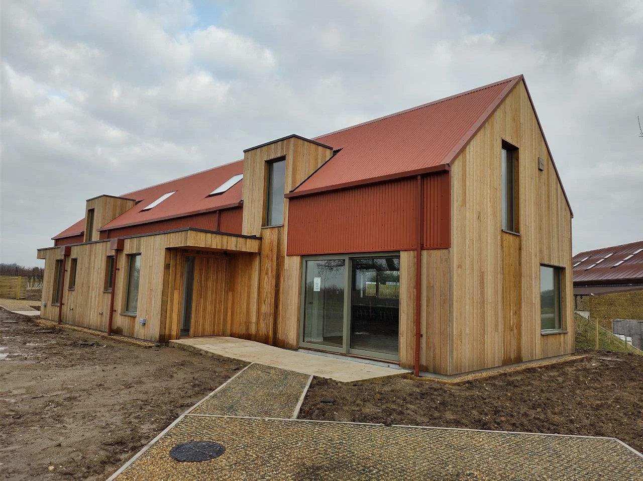 Newly constructed modern wooden house with a red metal roof and multiple windows, including a sliding glass door, on overcast day.