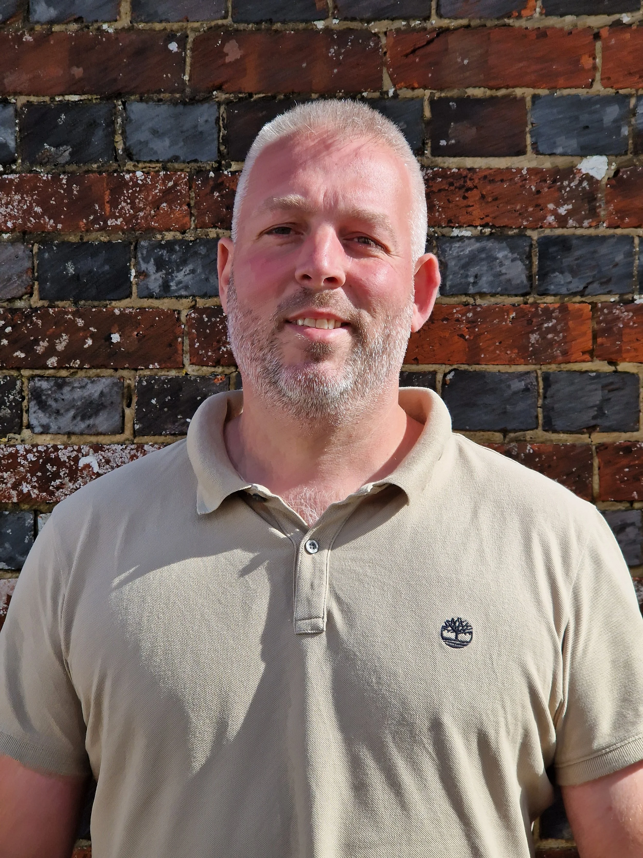 A man with short gray hair and a beard standing outdoors in front of a brick wall, wearing a beige polo shirt with a small logo on the left side.