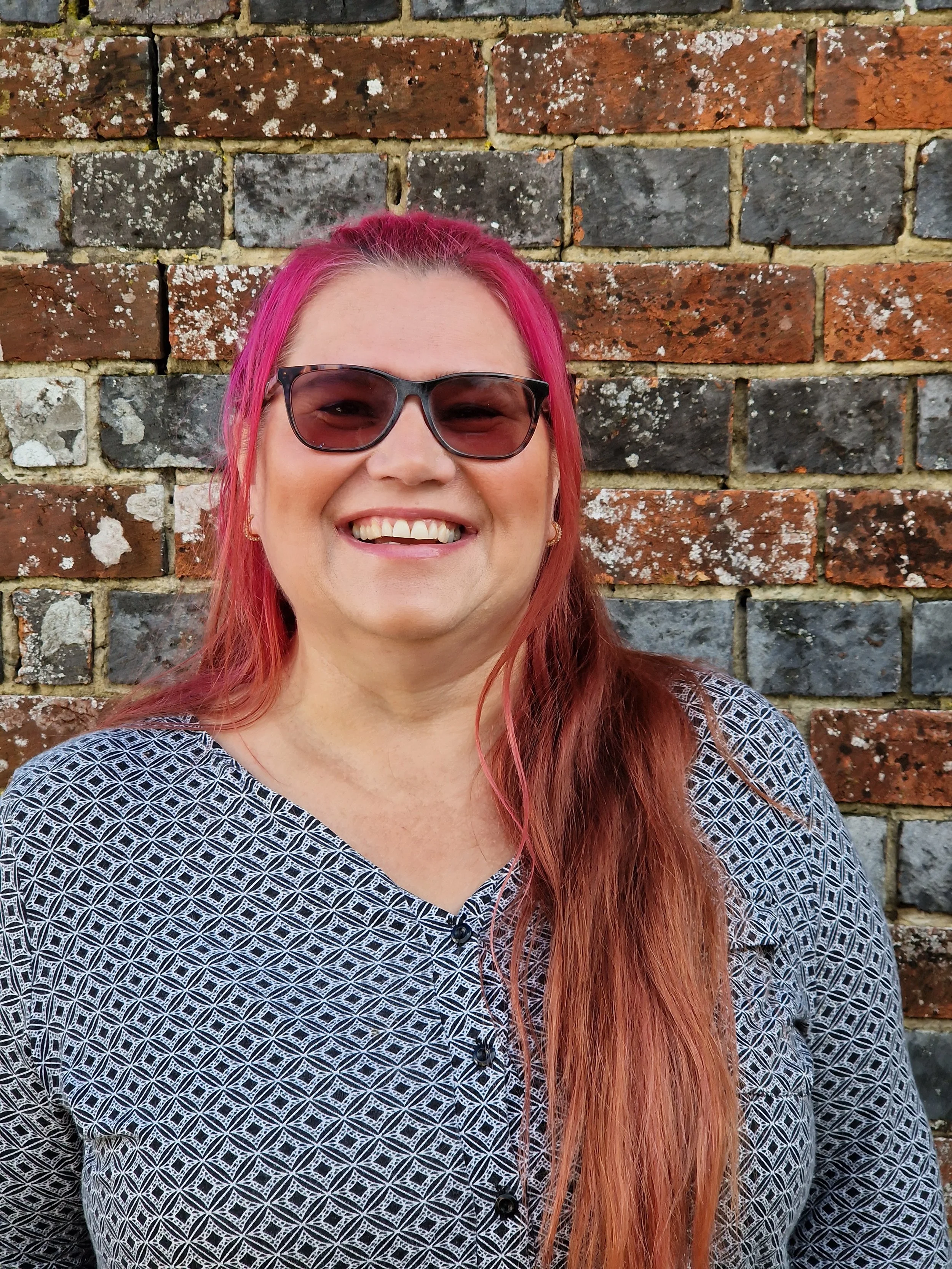 A woman with long pinkish-red hair wearing sunglasses and a patterned black and white top standing in front of a brick wall.