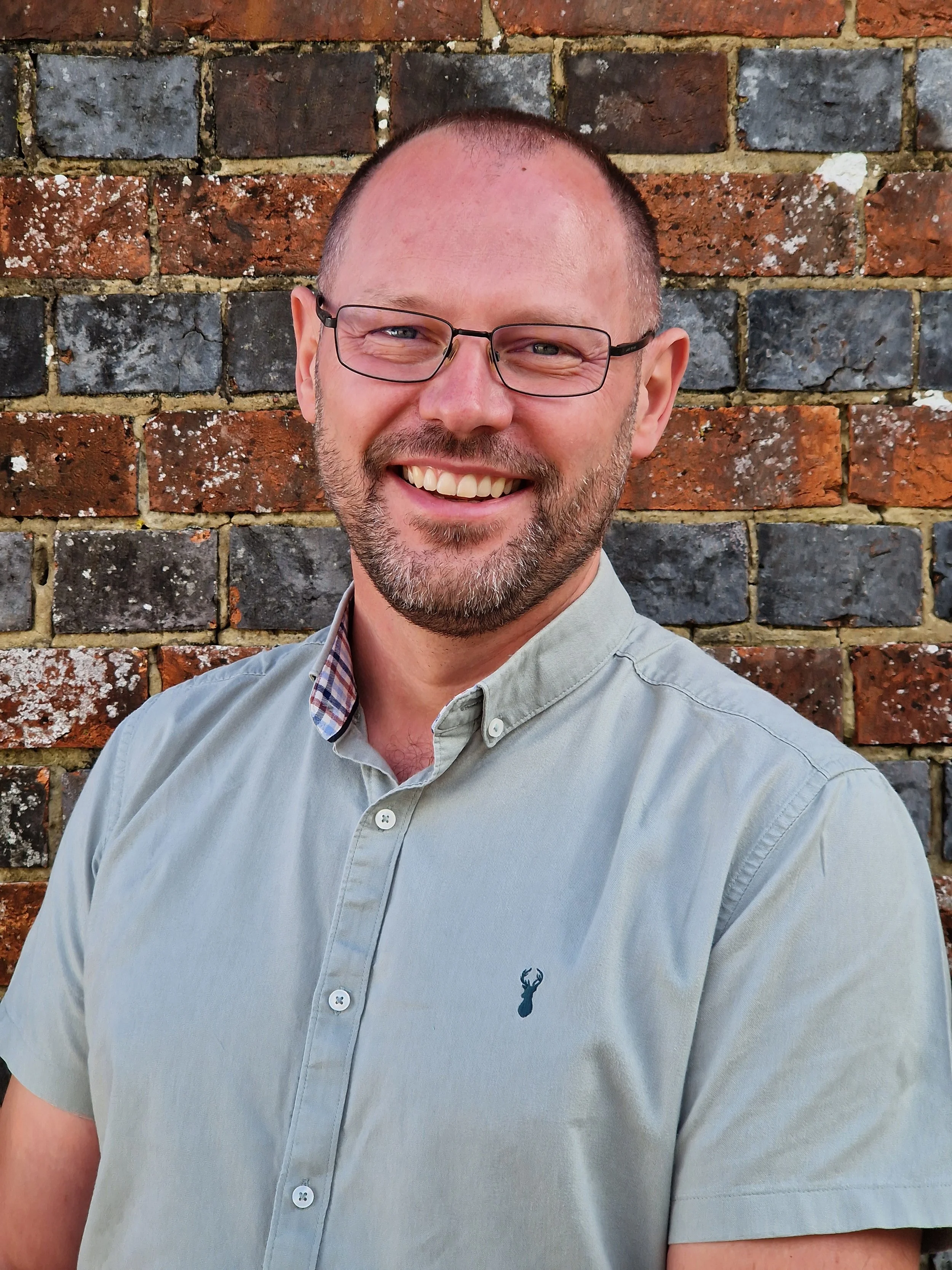 A smiling man with glasses, a beard, and short hair, wearing a light gray button-up shirt with a small navy emblem on the chest, standing against a brick wall background.