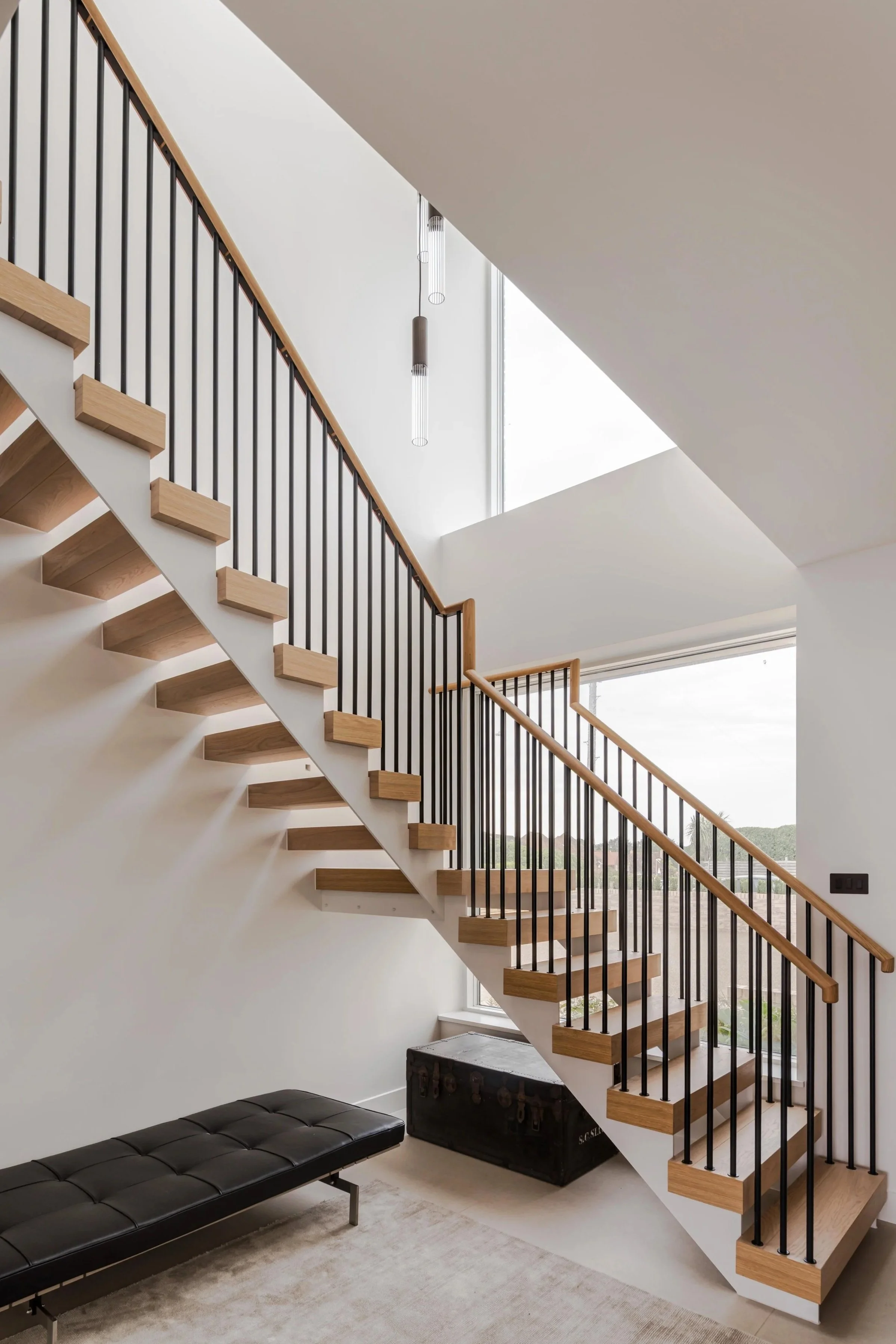 Interior view of a modern staircase with wooden steps, black metal railings, and a large skylight. There is a black leather bench and a vintage trunk beneath the staircase, with a window showing a partial view of the outdoors.