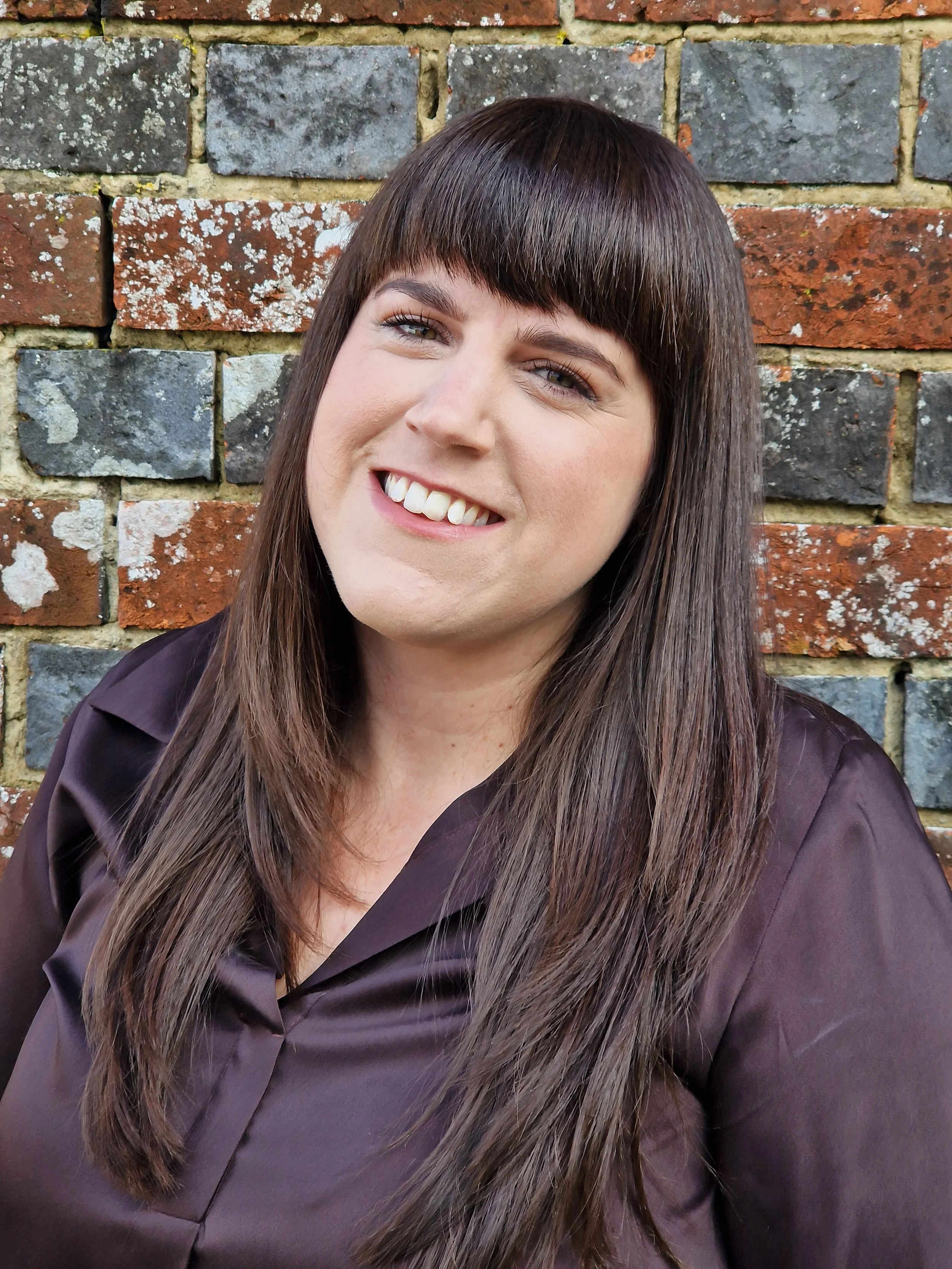 A woman with long brown hair and bangs smiling, wearing a dark brown blouse, standing in front of a brick wall.