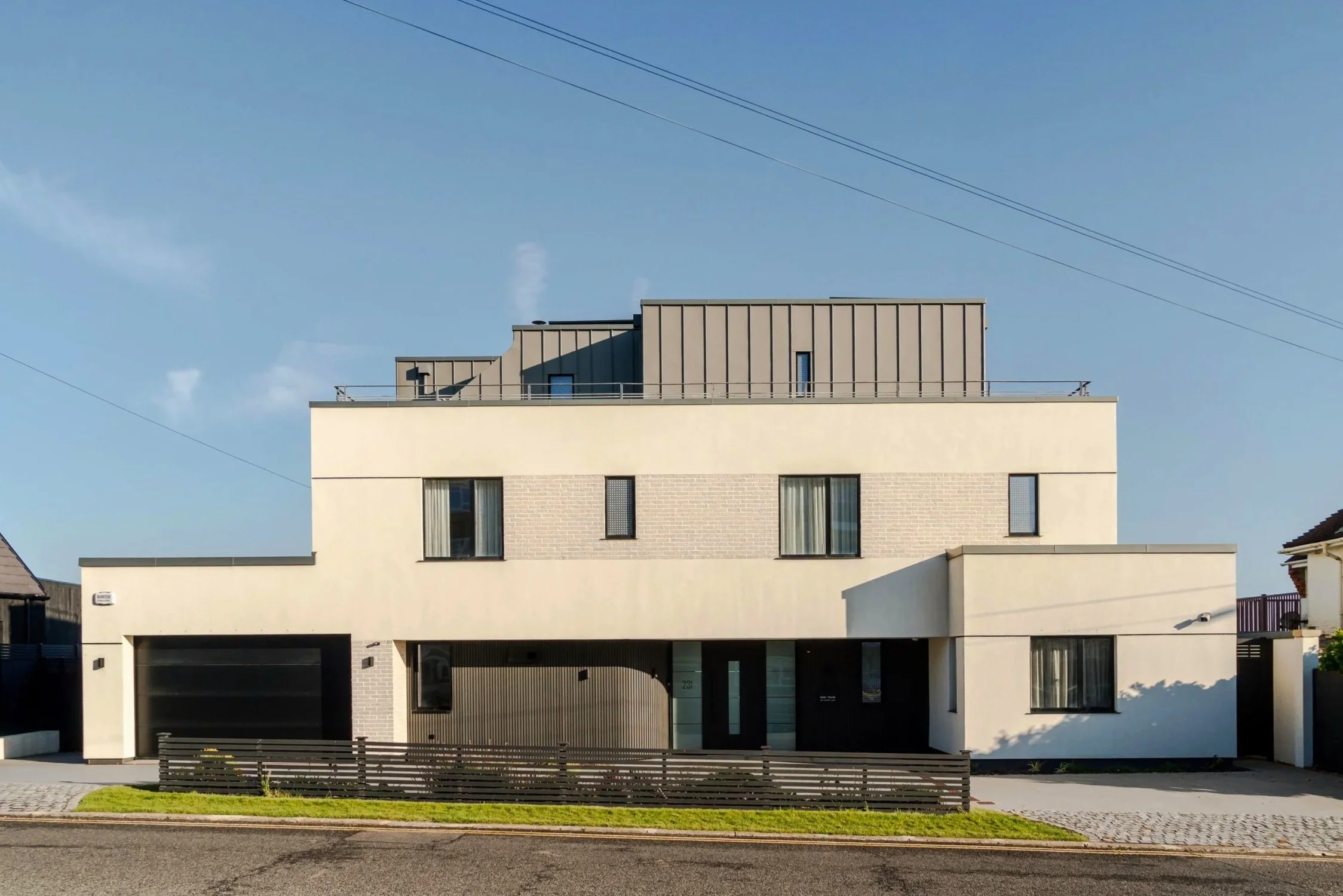 Modern multi-story residential building with a white facade, large windows, and a balcony at the top. The building features a garage with a black door on the ground level, a small green lawn in front, and is situated next to a sidewalk and street. Clear blue sky with power lines overhead.