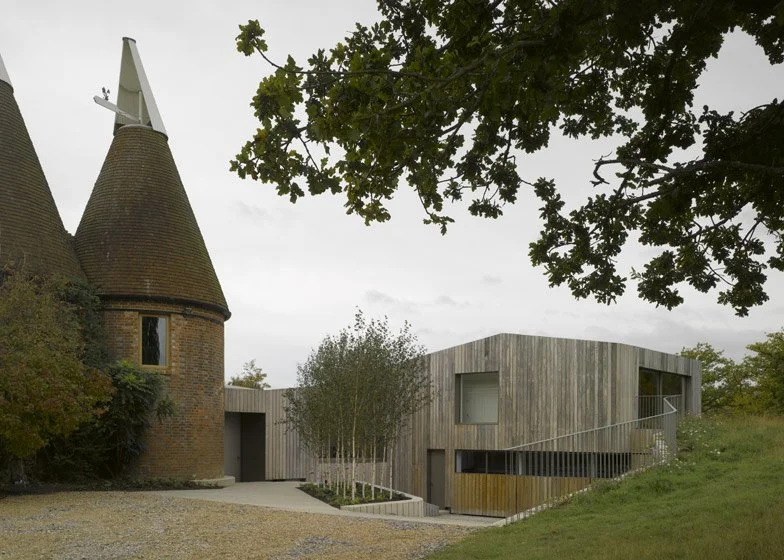 A historic brick turreted building with conical roofs next to a modern wooden and concrete structure, surrounded by trees and a gravel pathway, under a cloudy sky.