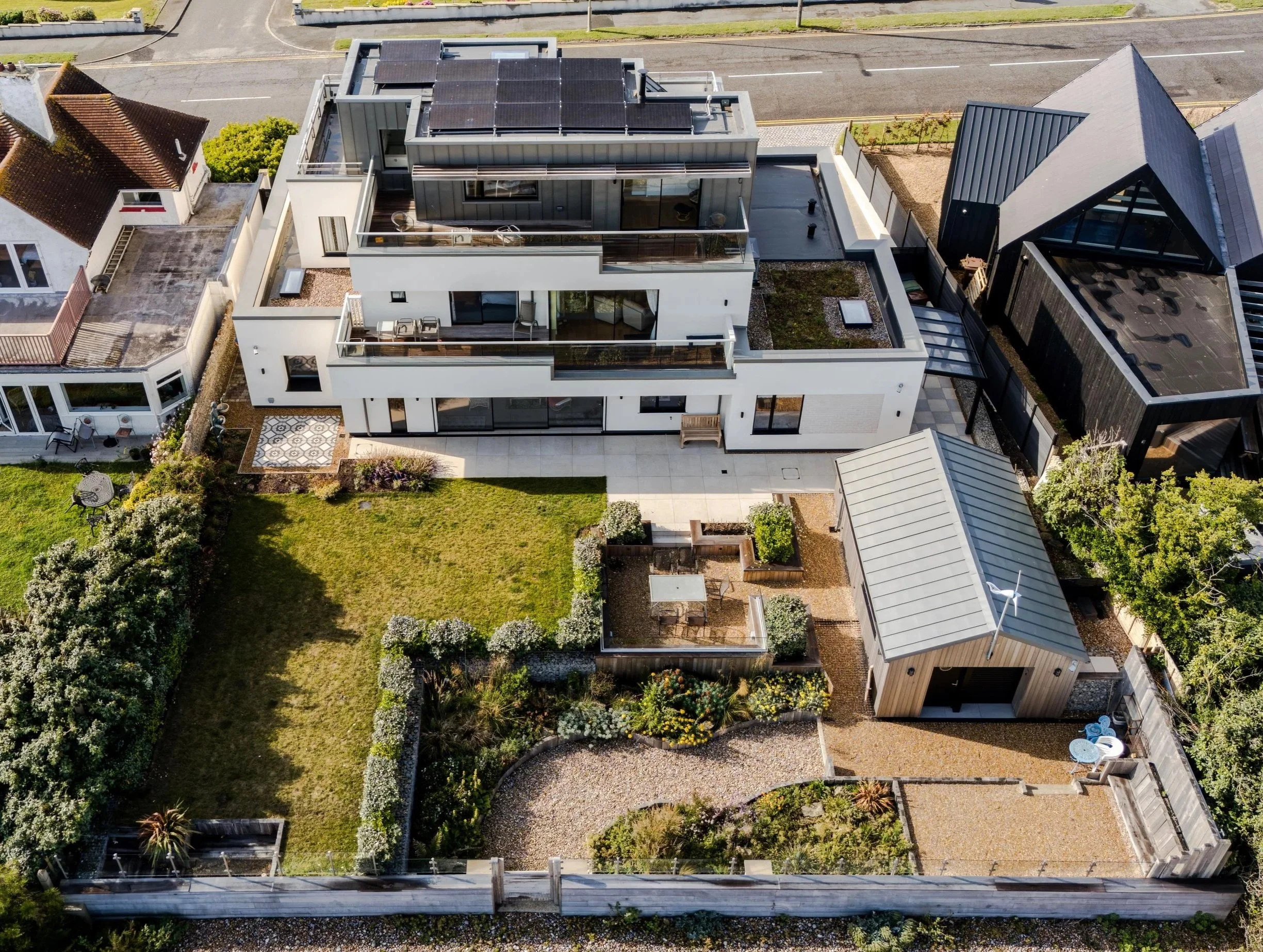 Aerial view of a modern white residential building with multiple balconies, solar panels on the roof, surrounded by a garden, neighboring houses, and a street.