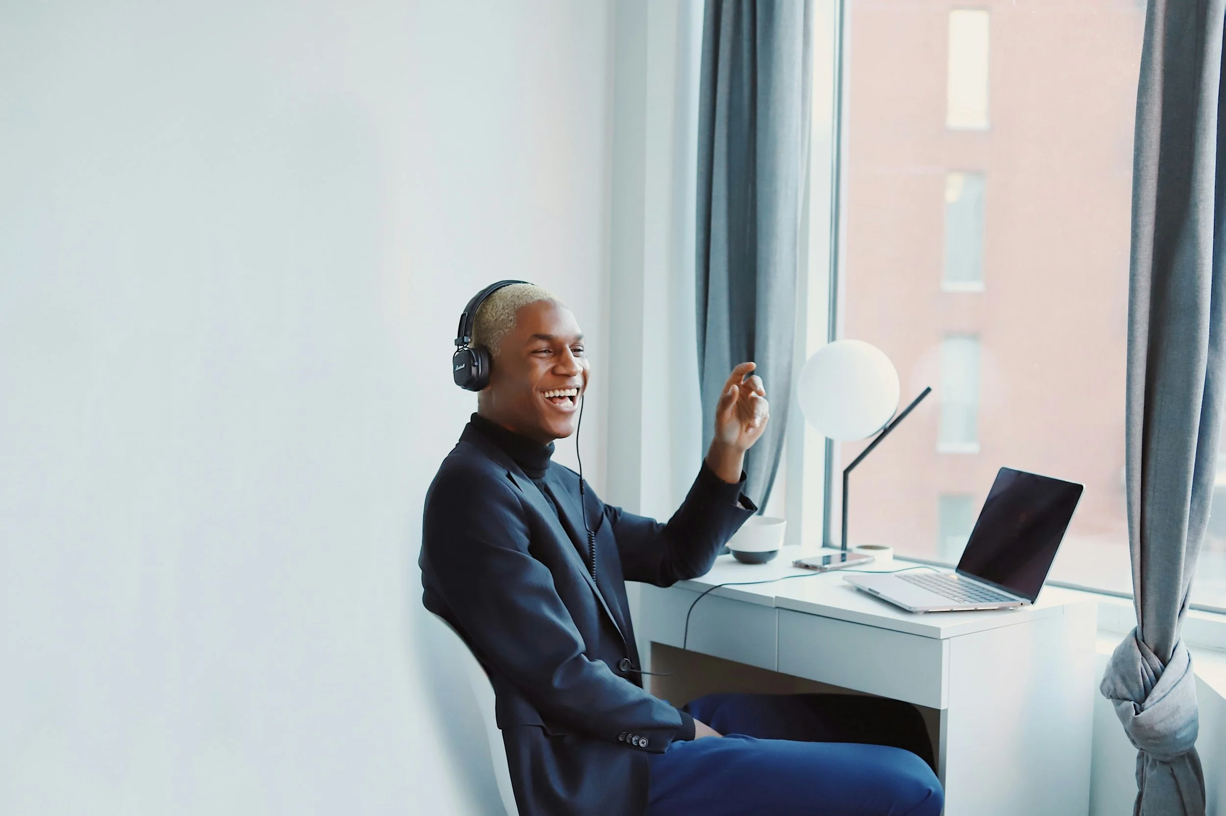 A young man with blonde hair and dark skin sitting at a white desk, wearing headphones, smiling and waving, with a laptop and a modern lamp on the desk, near a large window with curtains, in a bright room.