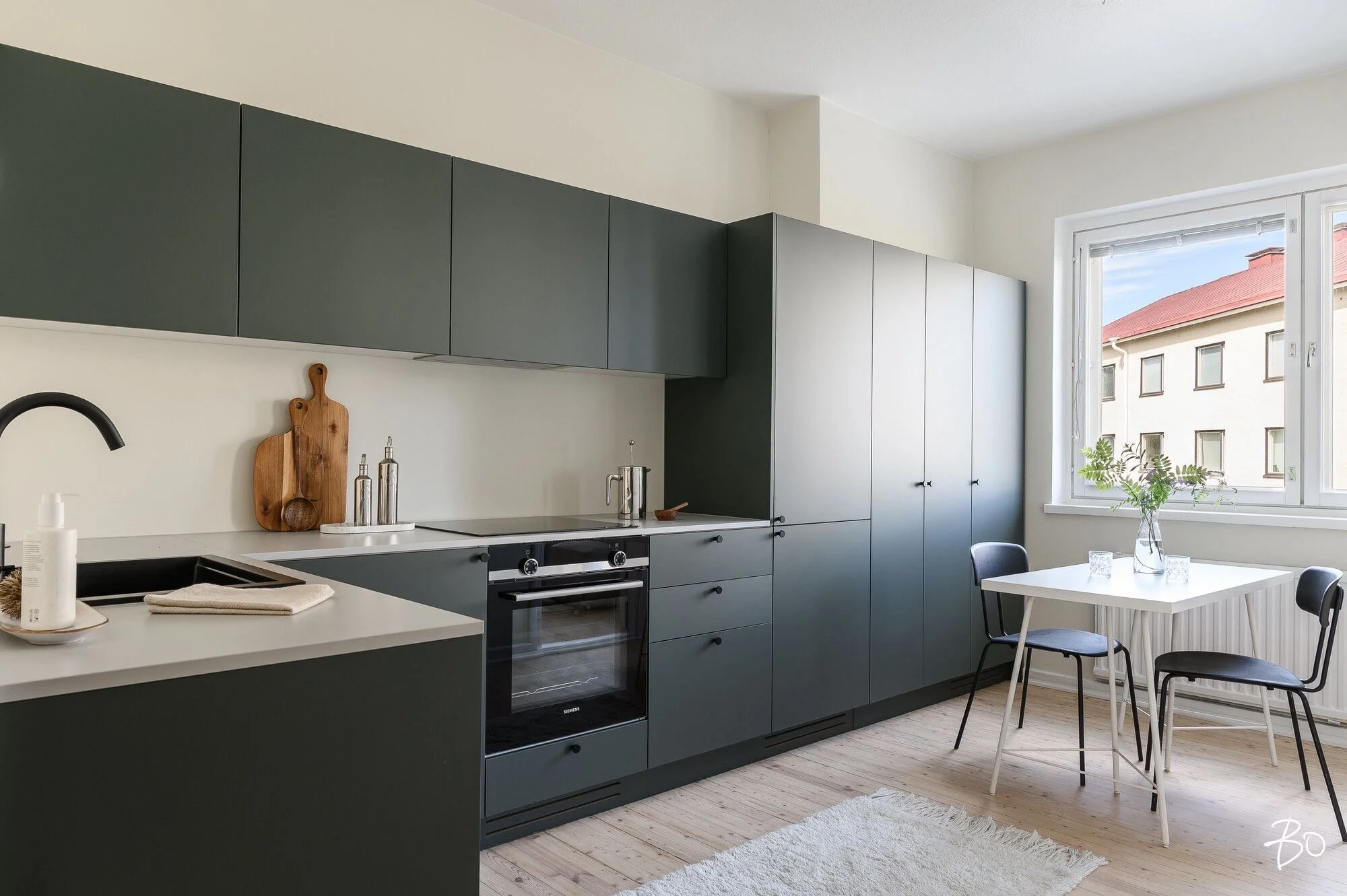 Tyhjä keittiö, jossa vihreät ylä- ja alakaapit, vaaleanharmaa työtaso ja pieni ruokailuryhmä ikkunan vieressä. Luonto näkyy ulkona ikkunasta.