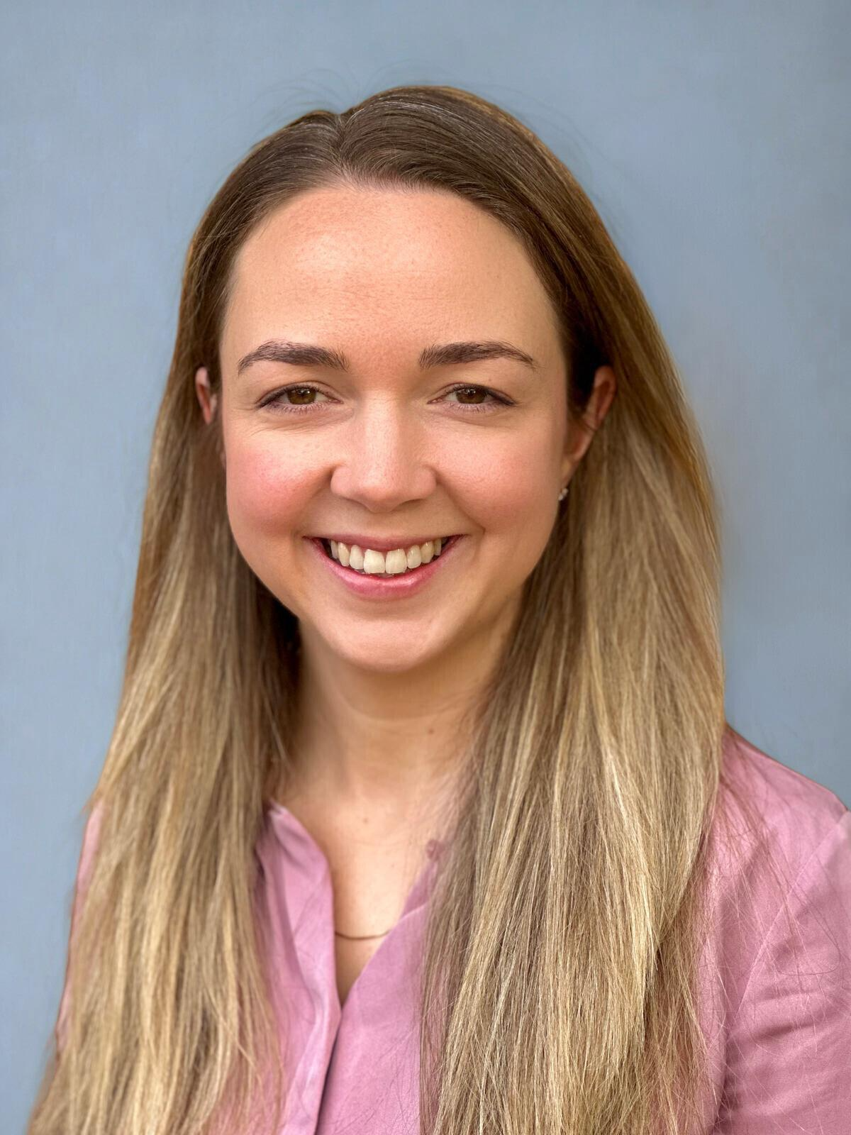 A young woman with long, light brown hair smiling in front of a plain blue background.
