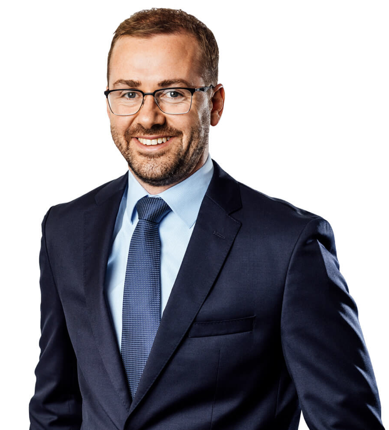 Portrait of a man with short brown hair and glasses, wearing a navy suit, light blue shirt, and blue tie, smiling against a white background.