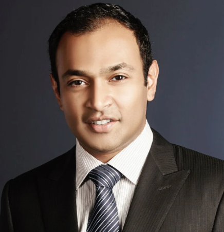 Professional man wearing a dark suit, white shirt, and striped tie, smiling against a dark background.