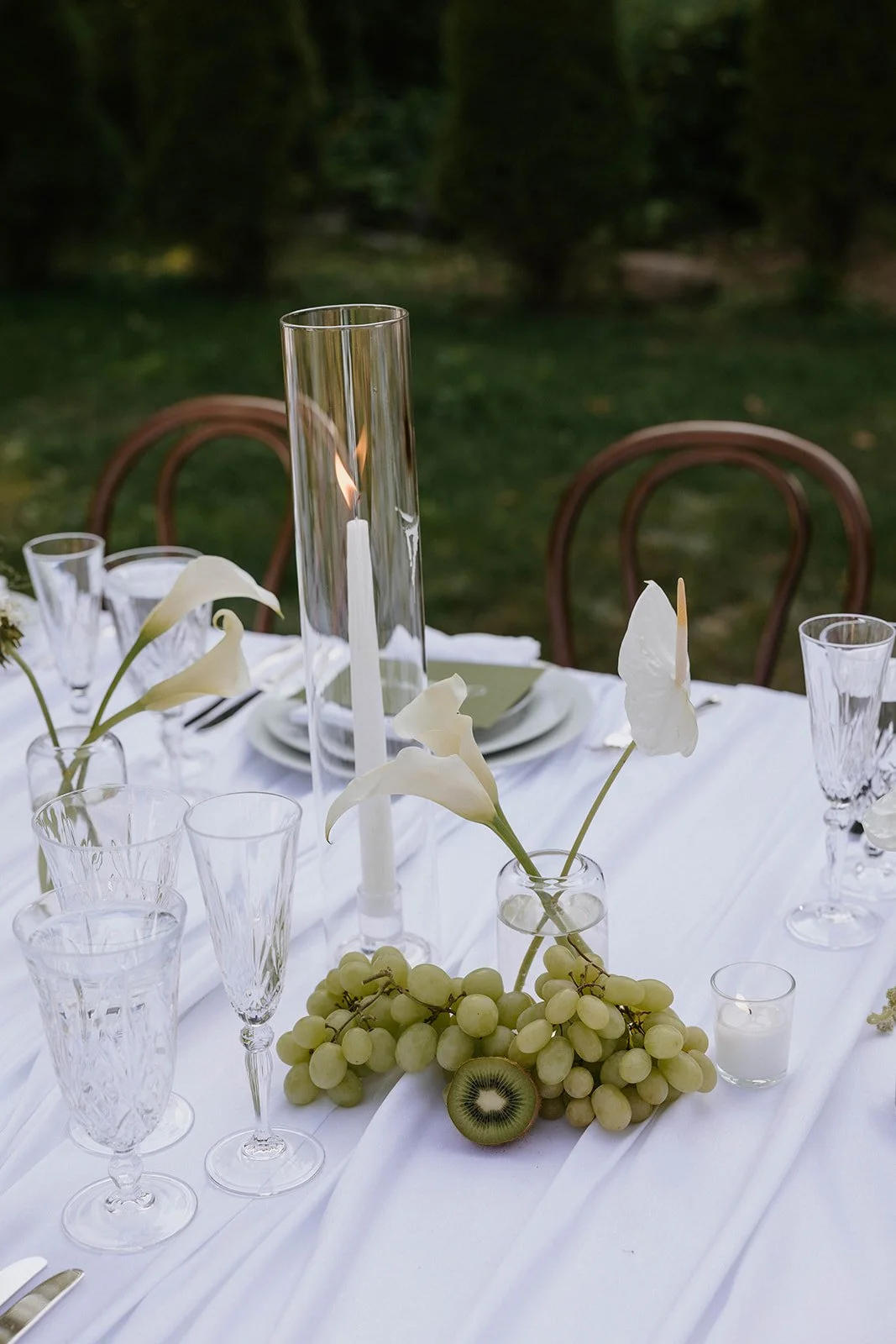 Elegant outdoor table setting with white flowers, grapes, a kiwi, glassware, candles, and a tall glass candle holder with a lit candle, against a green garden background.