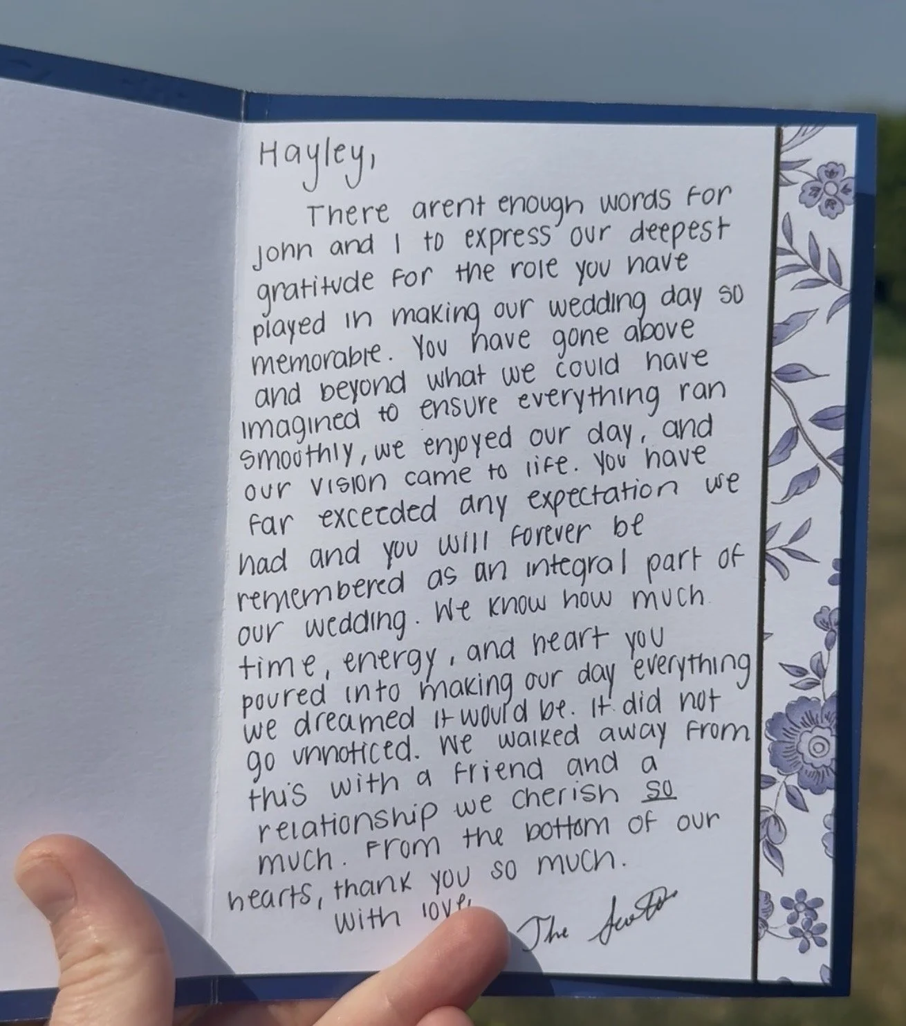 Inside view of a handwritten wedding thank you card, with purple floral border on the right side, being held by a person's left hand outdoors.