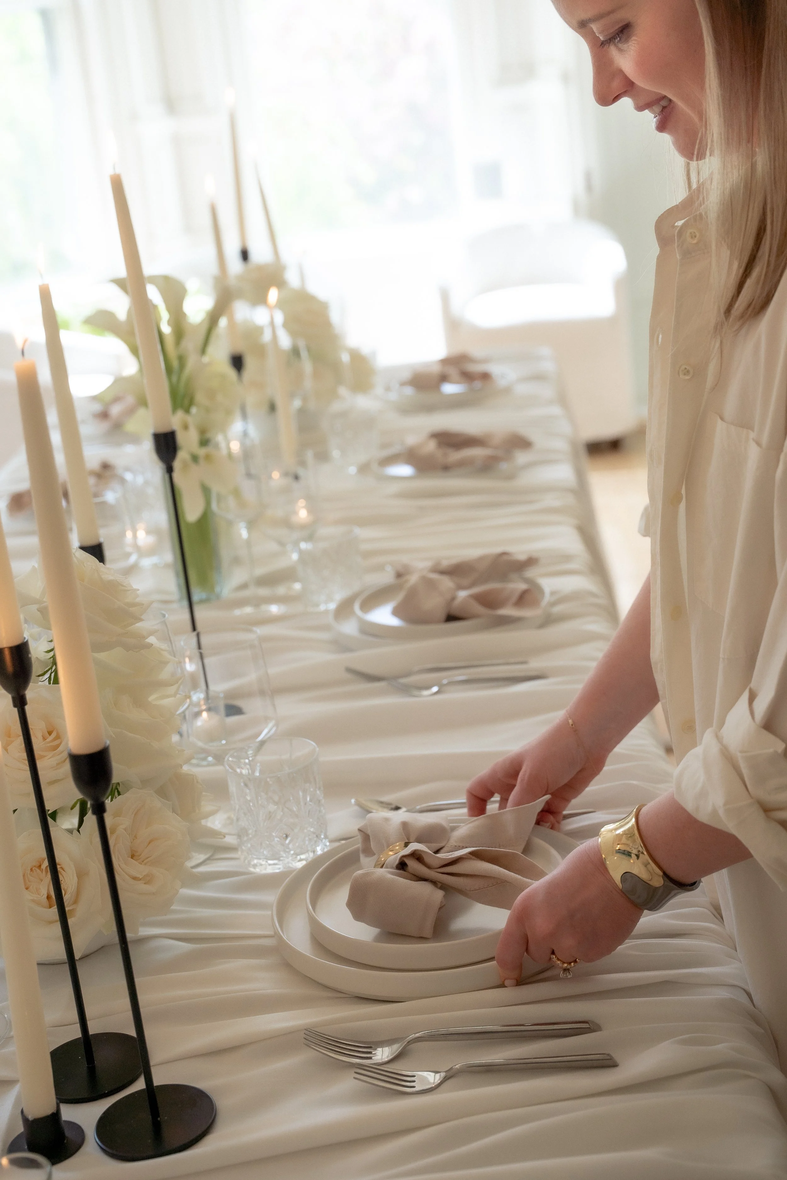 Wedding styling executed by Hayley, you see her here arranging place settings at a formal table decorated with white flowers and candles, in a bright room with natural light.