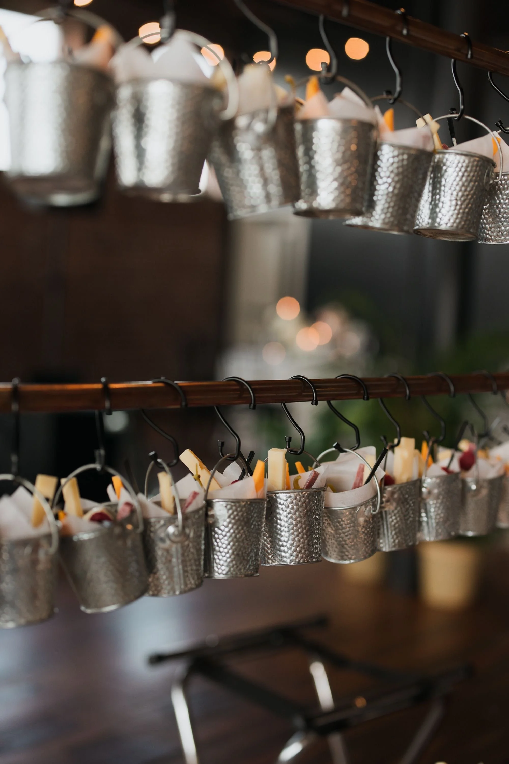 Metal buckets hanging from hooks on a shelf, filled with assorted cheeses and garnishes, with blurred warm lighting in the background.