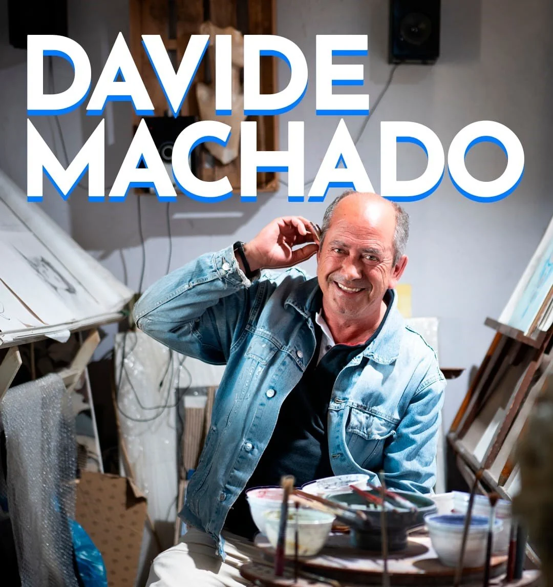 A man smiling and wearing a denim jacket, sitting at an art studio cluttered with painting supplies and artworks, with large text overlay of "DAVIDE MACHADO".