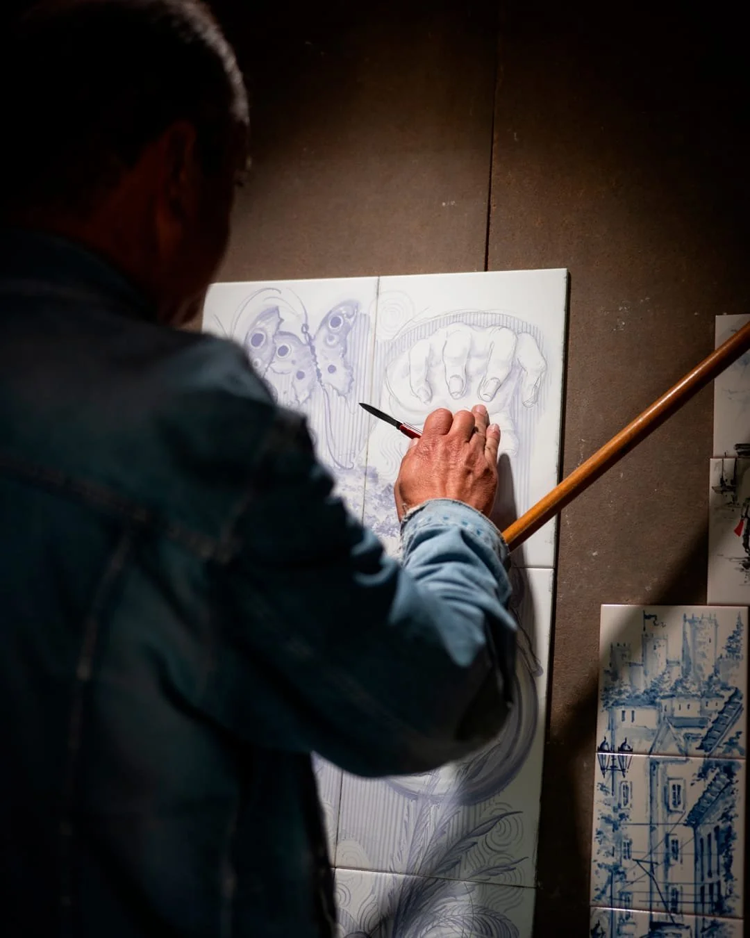An artist sketches on a large canvas, depicting a butterfly and a hand, in an art studio with ceramic tiles and drawings on the wall.