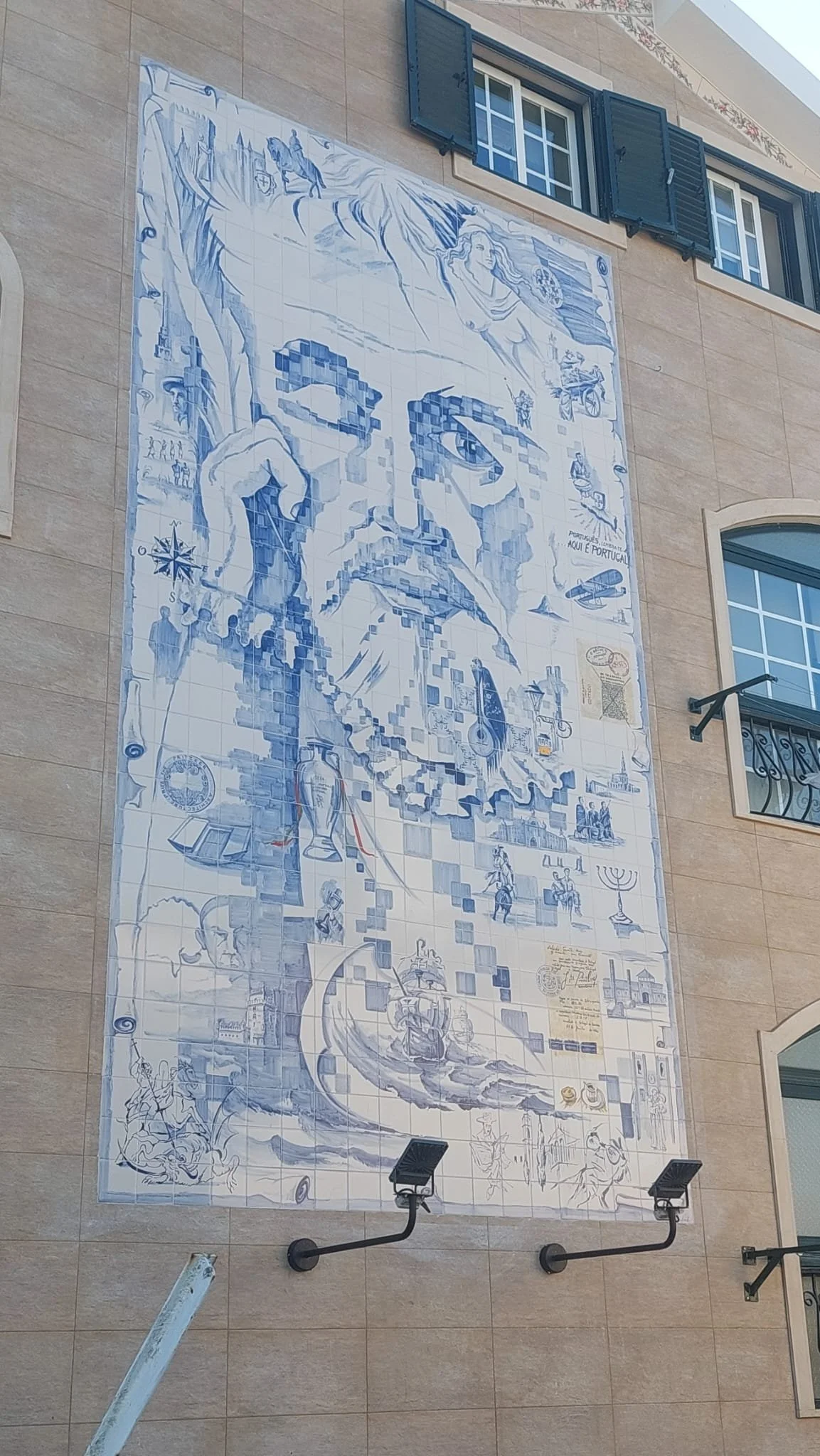 Large blue and white mural with a pixelated portrait of a man in the center, surrounded by various cultural and historical images, including a woman, a ship, and native symbols, on the exterior of a building.