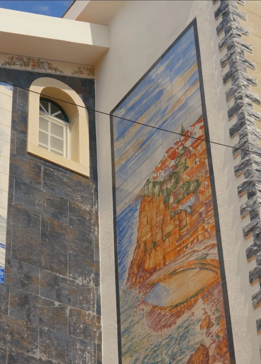 Large mural on the side of a building depicting a coastal scene with a cliffside village, water, and sky. Adjacent to the mural is a beige wall with a window and decorative stone trim.