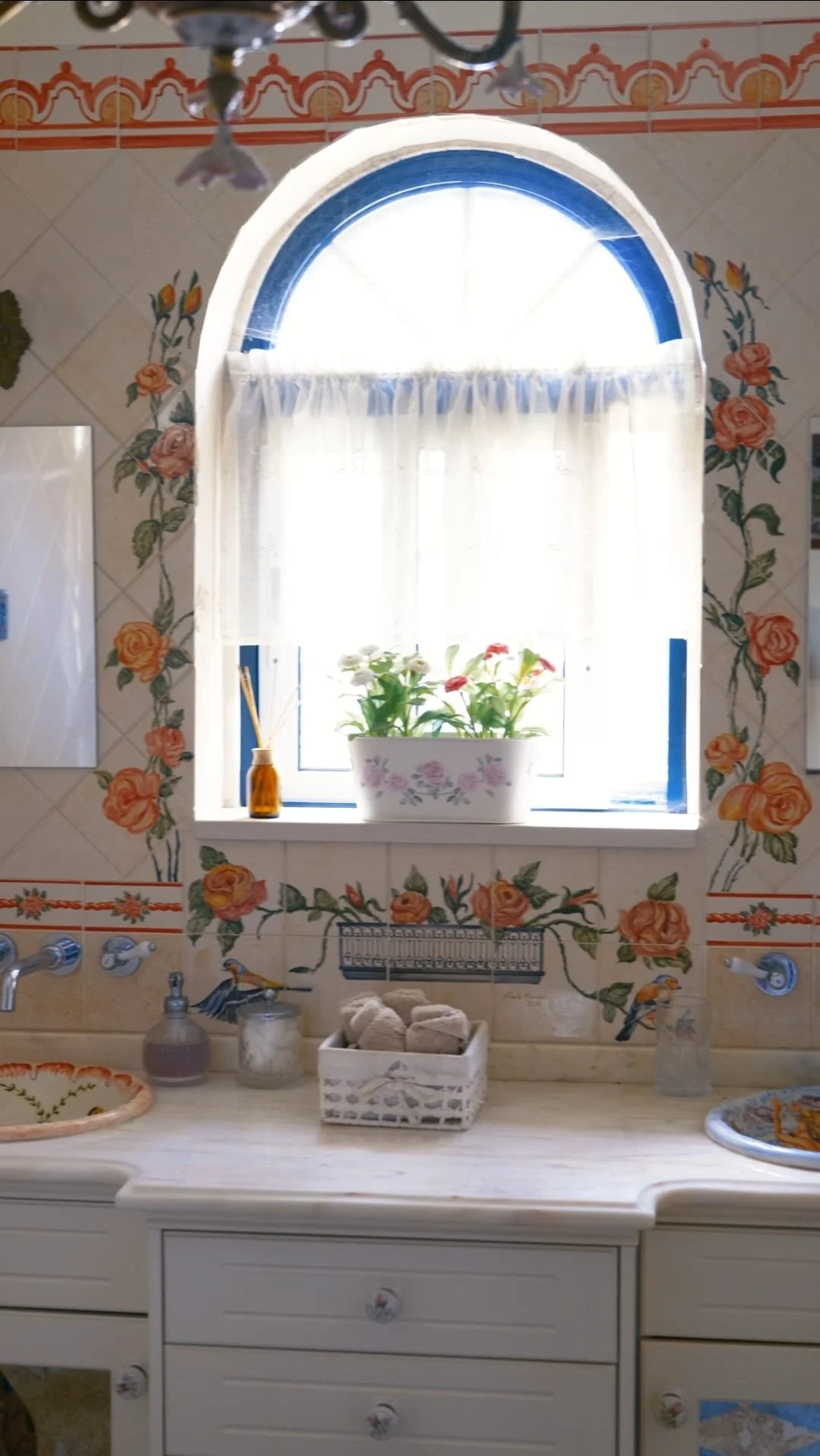 Kitchen window with floral and bird decor, a white cabinet counter with soap, sponge, and decorative dishes, and a potted plant on the window sill.