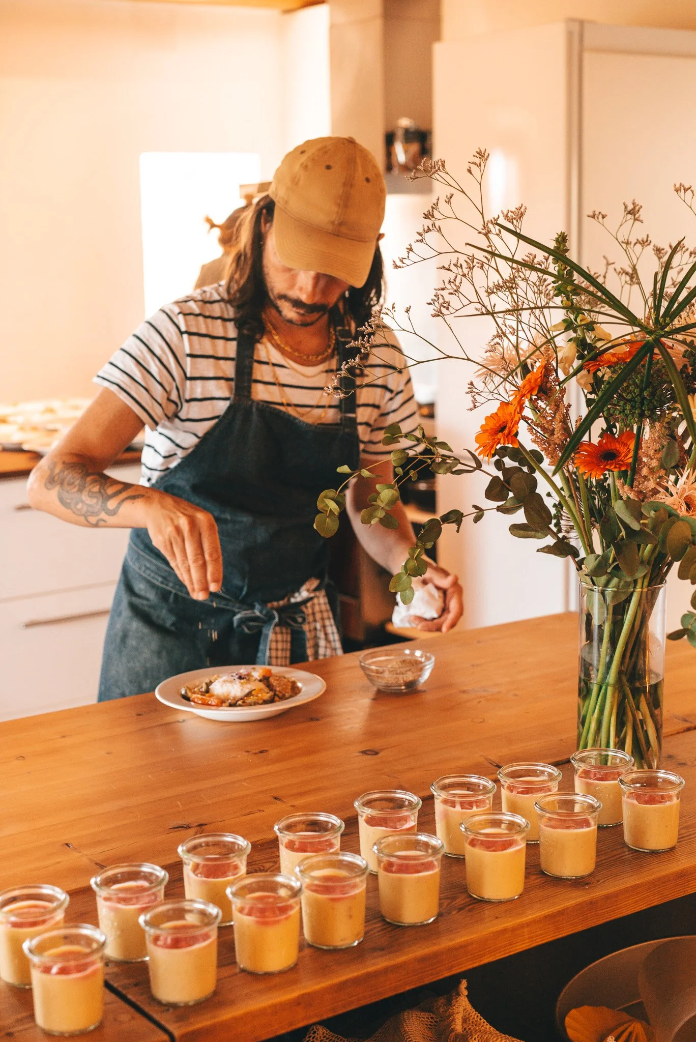 Una persona sirviendo comida en una cocina, con varios frascos de postre en un mesón de madera y un ramo de flores en un florero al frente.