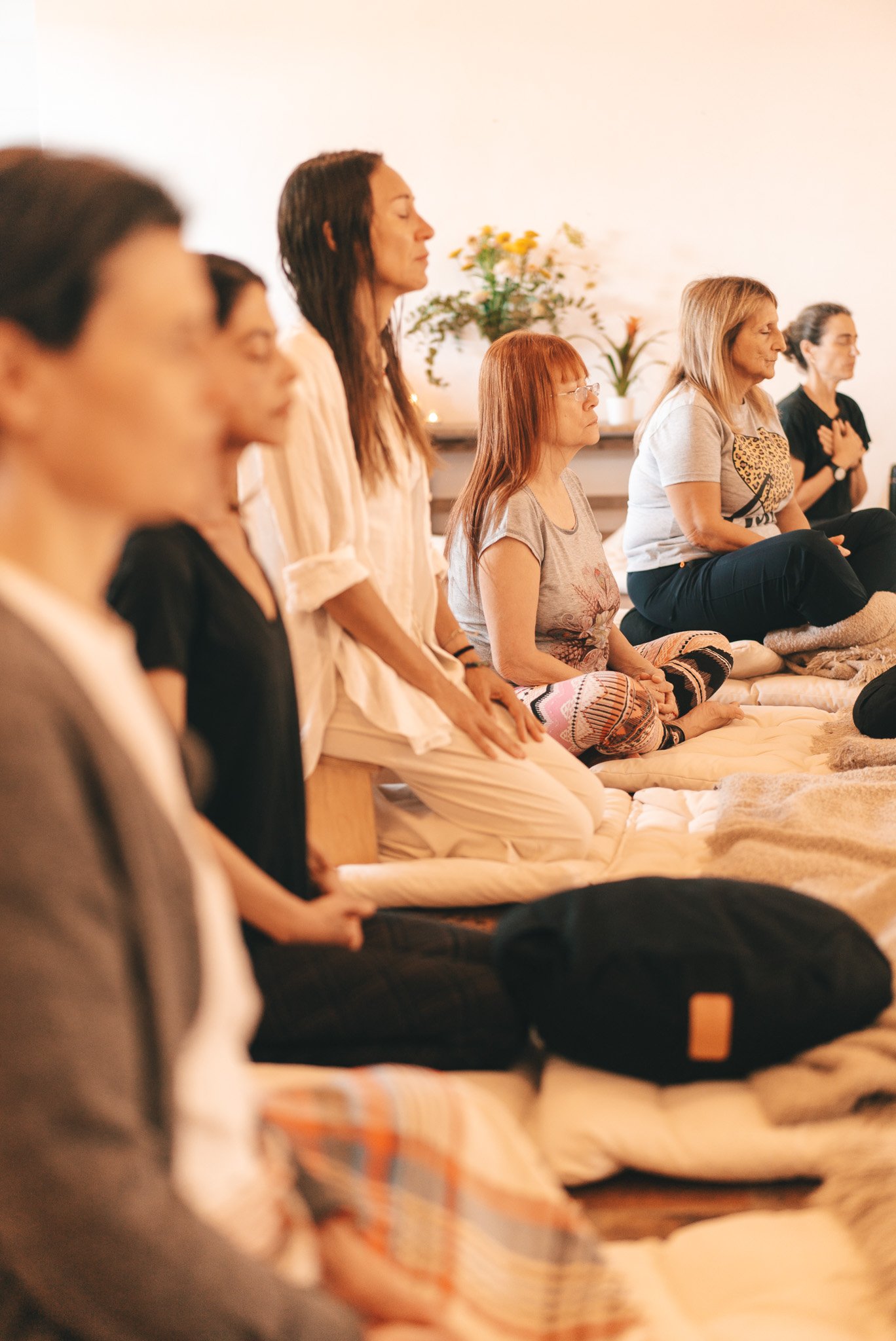 Varias mujeres en posición de meditación en una sala con plantas y flores.