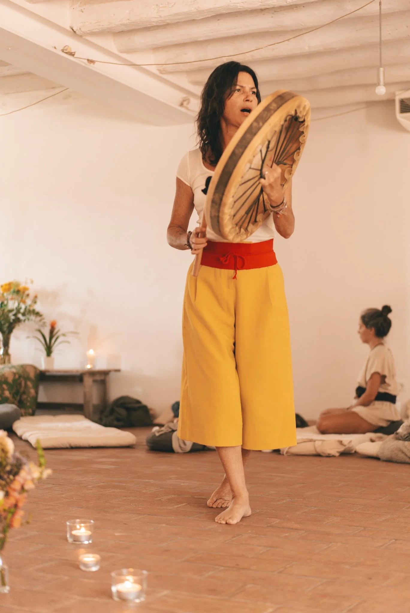 Mujer tocando un tambor, de pie, en una habitación con decoraciones suaves y gente en el fondo.