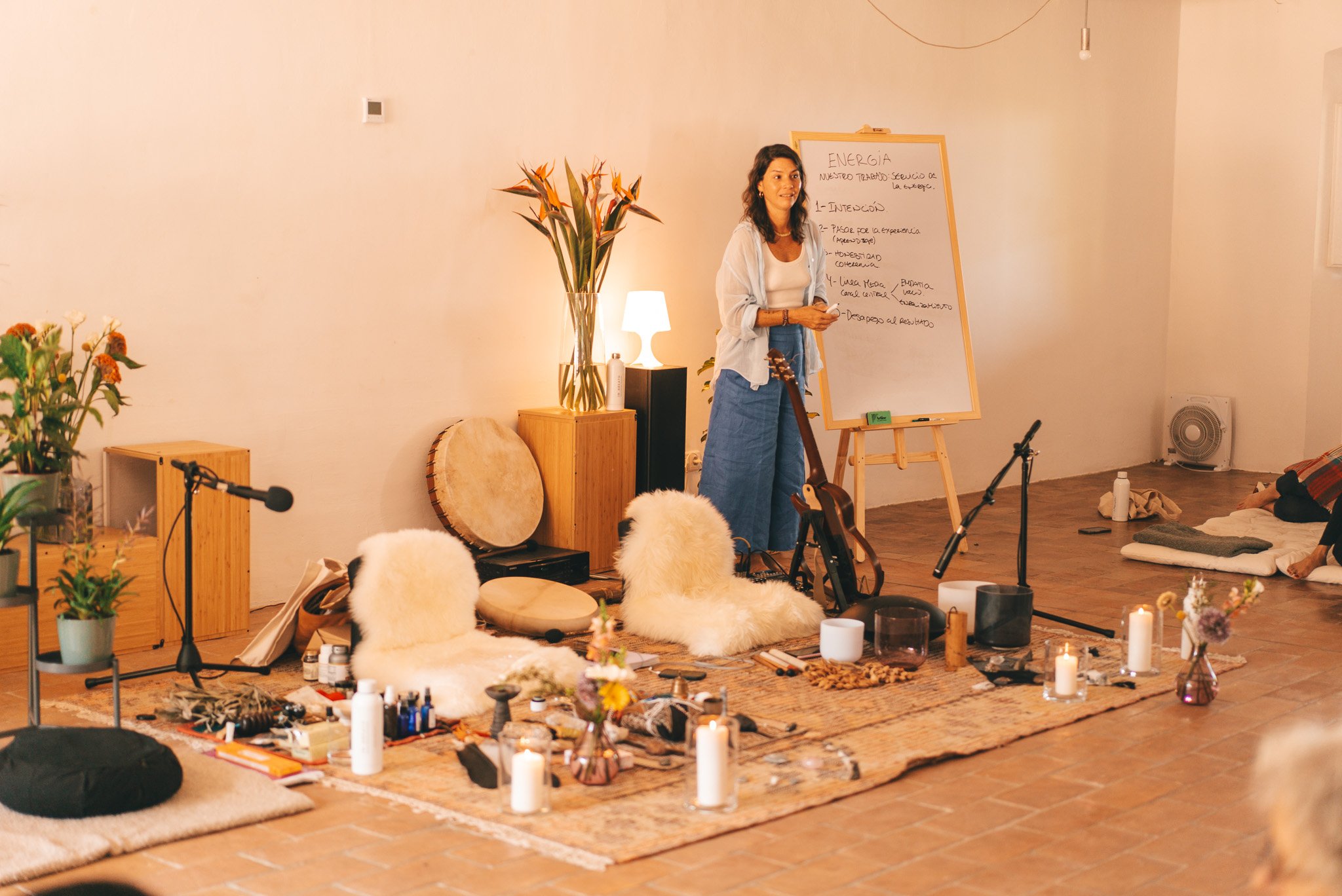 Mujer dando una charla en un espacio decorado con plantas, velas y instrumentos musicales, con una pizarra y un micrófono en el escenario.