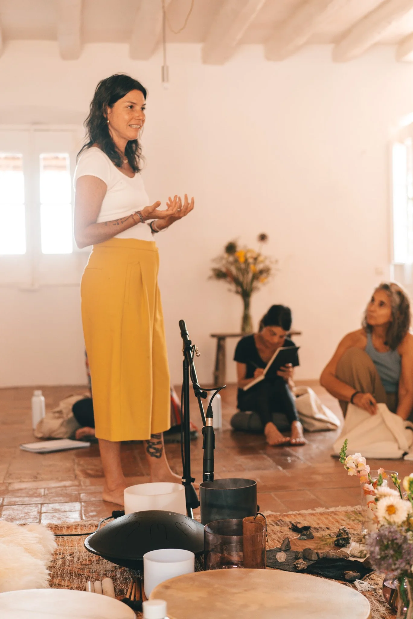 Mujer de cabello rizado y blusa blanca, de pie, hablando a un grupo en un ambiente calmado y decorado con flores y símbolos de armonía.