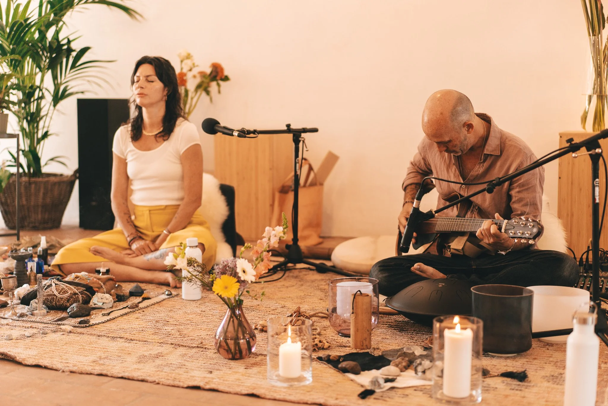 Una mujer y un hombre tocando instrumentos en una meditación o sesión musical espiritual en una habitación decorada con velas, flores y objetos de energía.