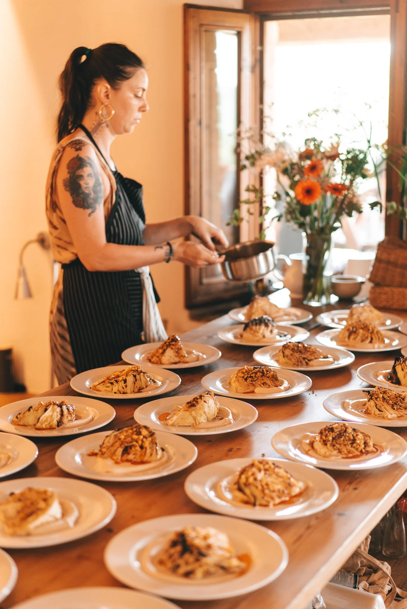 Una mujer sirviendo pastel en una mesa con varios platos, en un ambiente cálido y bien iluminado con flores en un jarrón.