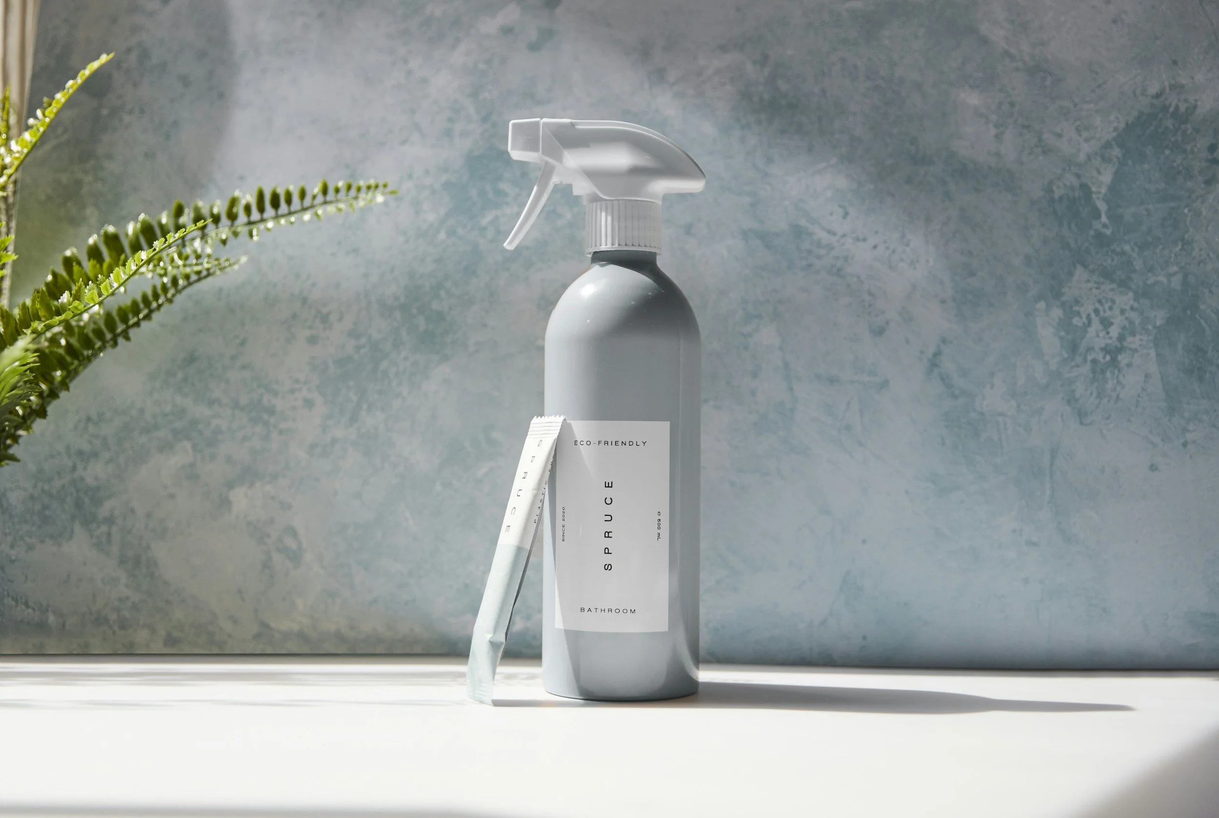 A gray spray bottle labeled 'Eco-Friendly' and 'Bathroom' next to a white package on a white surface with a textured blue wall background.