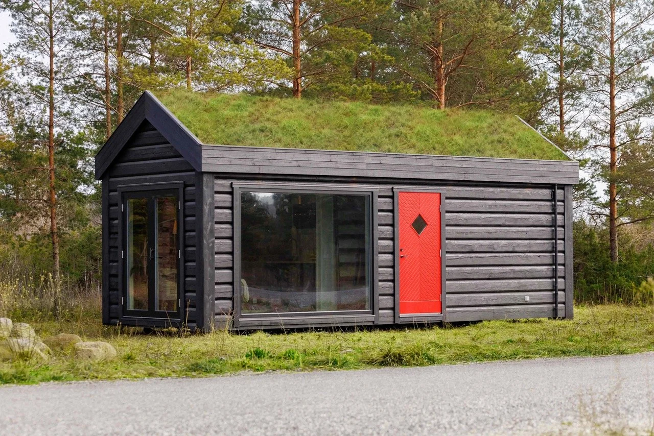 Small black cabin with a green grass and trees background, featuring large glass windows and a red front door with a diamond-shaped window.