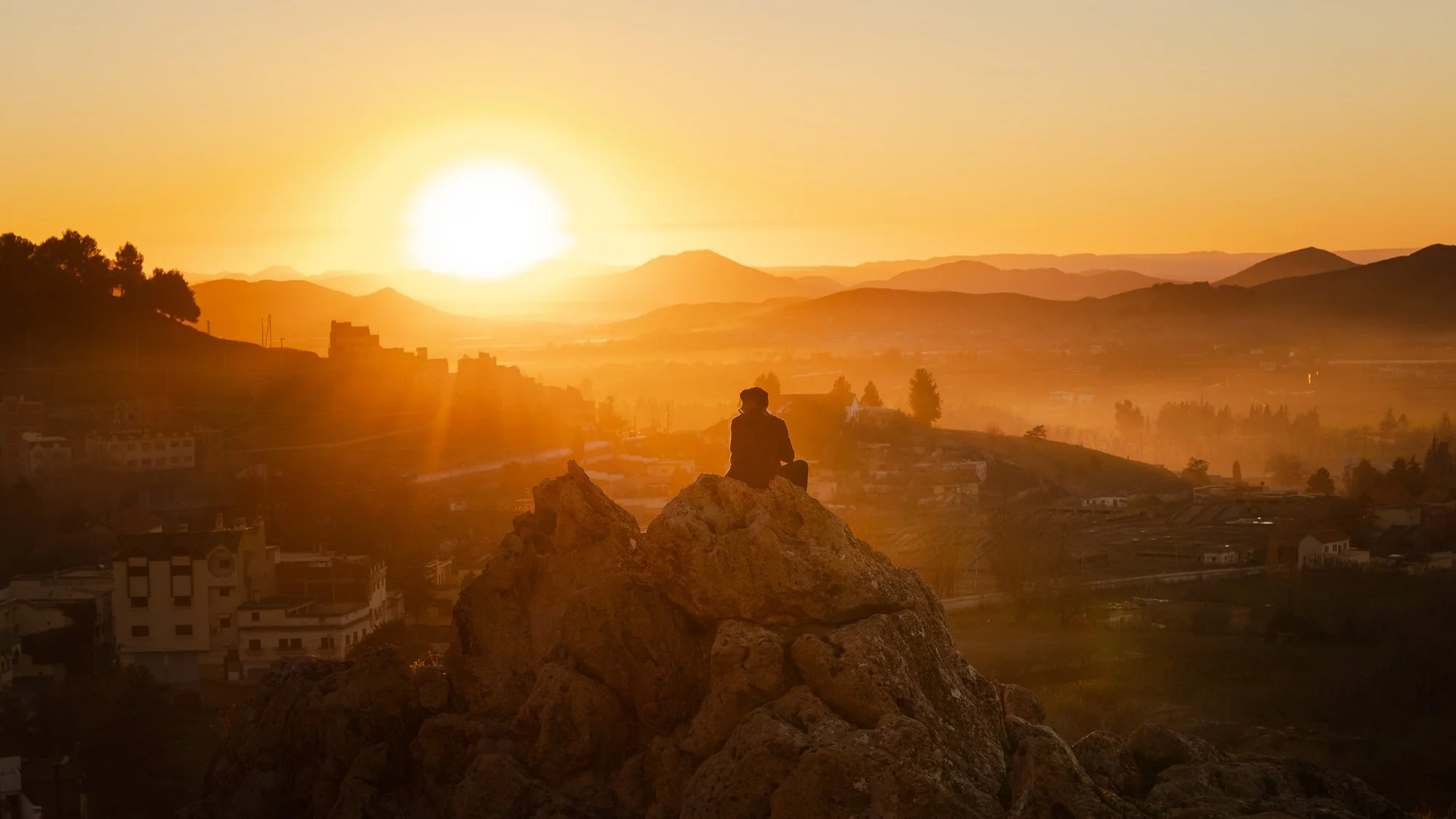 Eine Person sitzt auf einem Felsen, während die Sonne im Hintergrund untergeht, mit Bergen und einer Stadt im Tal.