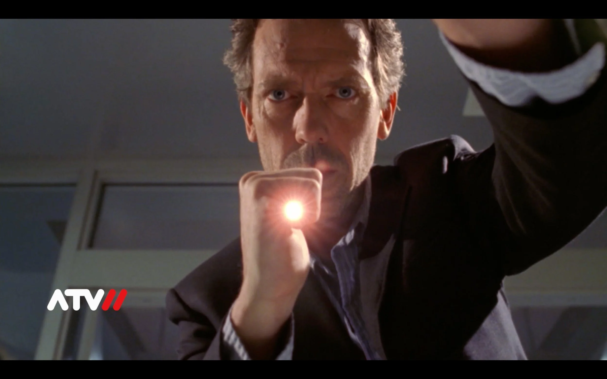 Ein Mann hält einen Taschenlampe, die direkt in die Kamera zeigt, während er nach unten schaut, mit einem ernsthaften Gesichtsausdruck.