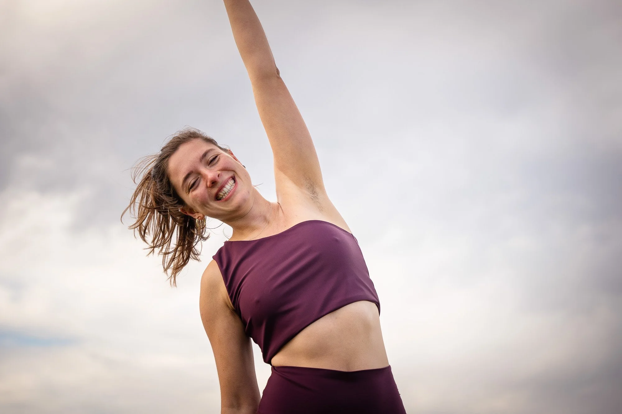 Eine lachende Frau mit braunem, schulterlangem Haar trägt eine dunkelviolette Sportbekleidung im Freien bei bewölktem Himmel.