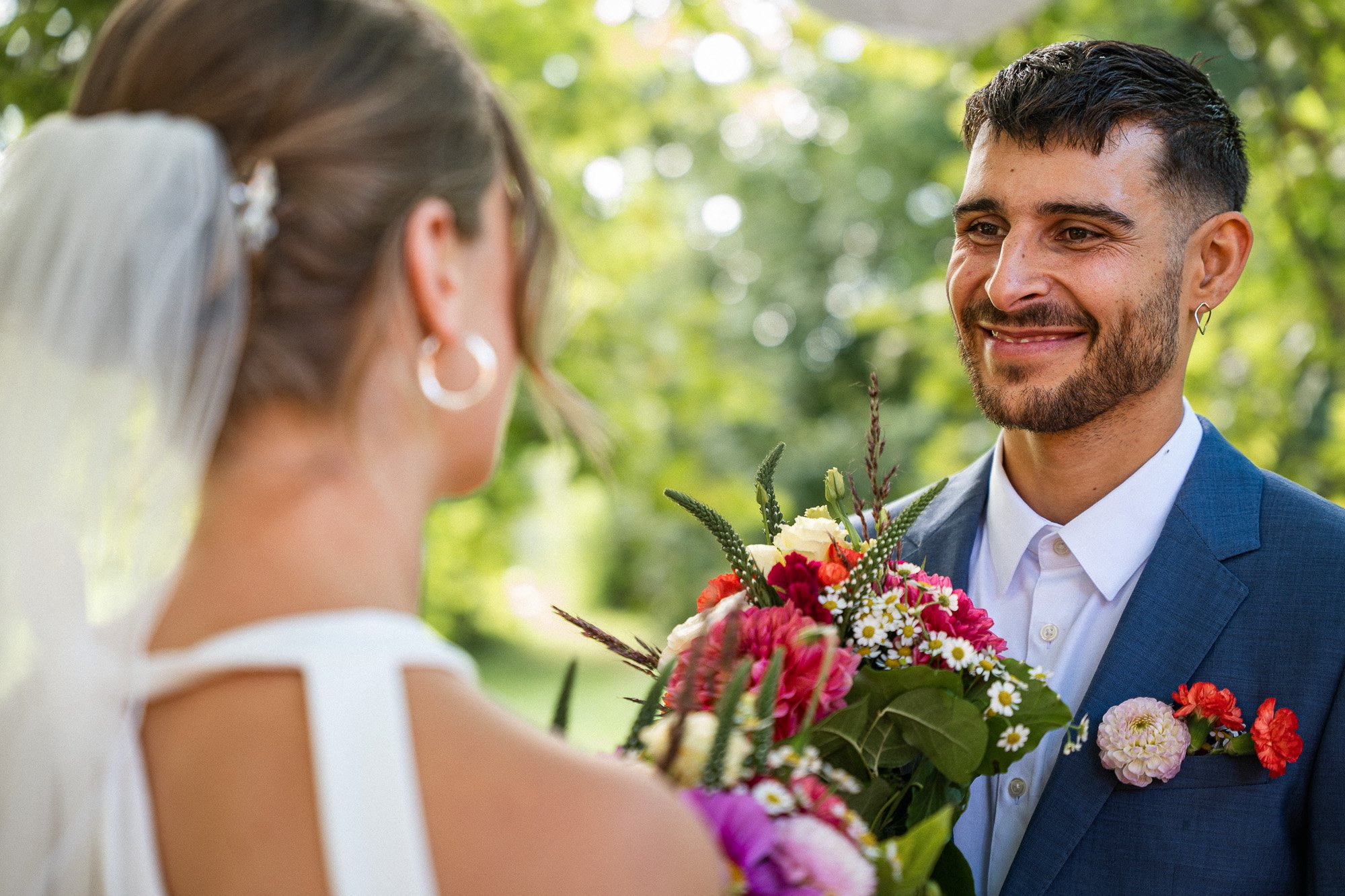 Ein Mann in einem blauen Anzug hält einem Brautpaar einen Blumenstrauß während einer Hochzeitszeremonie im Freien mit grünen Bäumen im Hintergrund.