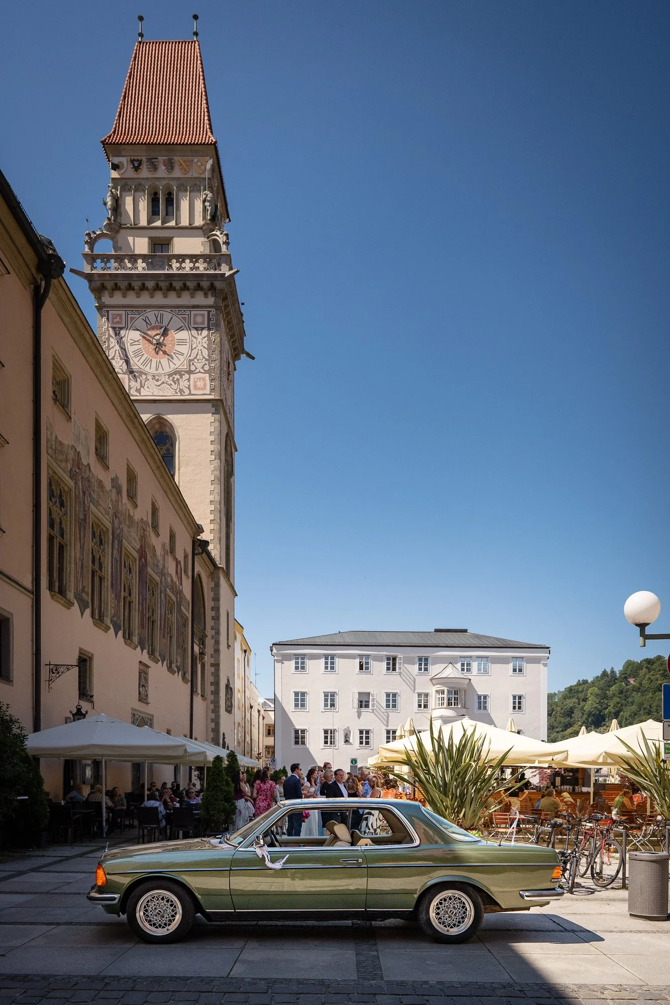 Historische Stadtstraße mit Uhrturm, Menschen, ein parkendes Oldtimer-Auto und Cafés im Freien bei sonnigem Wetter.