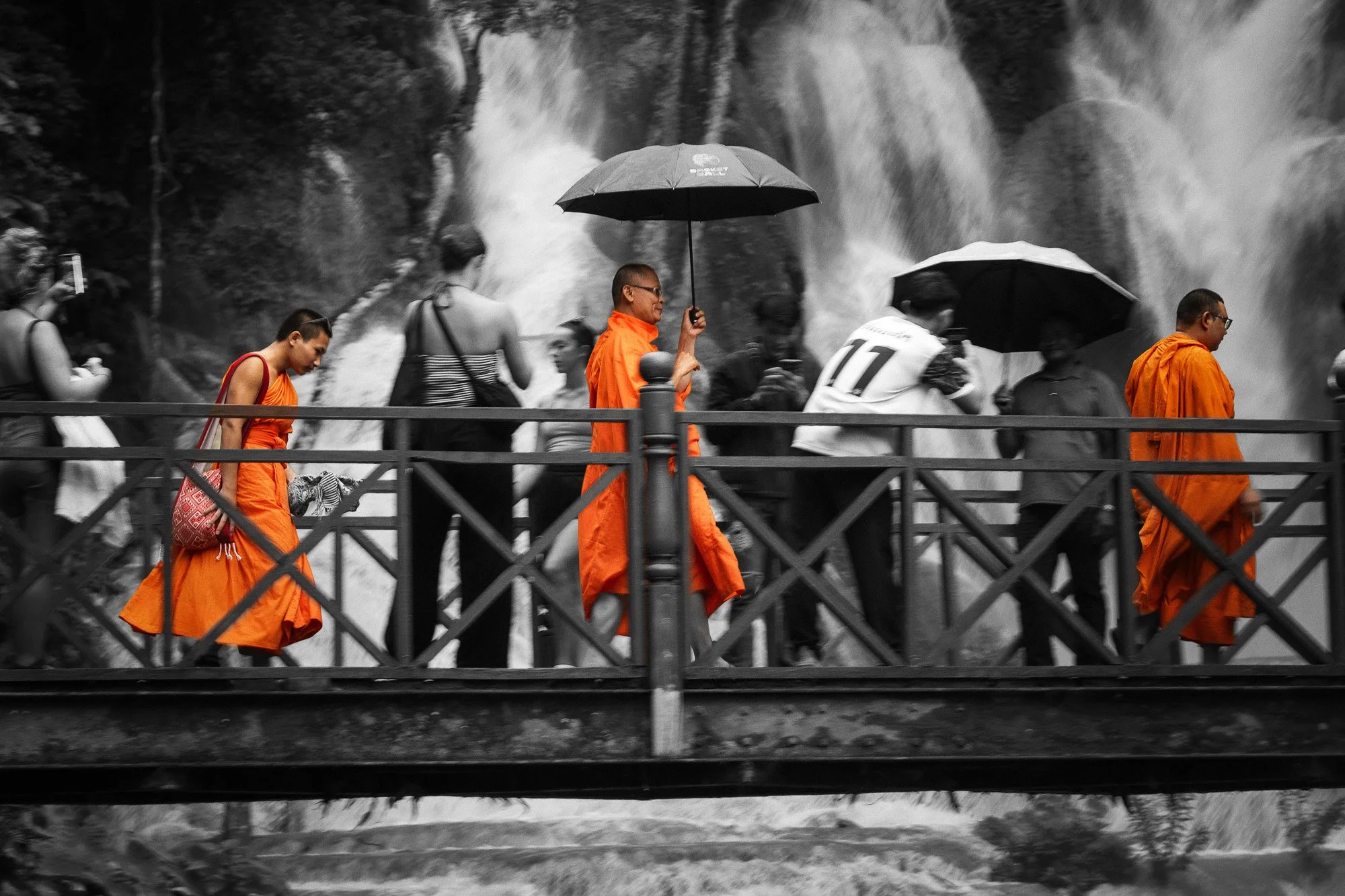 Menschen gehen auf einer Brücke über Wasserfall, einige tragen orange Roben, andere haben Regenschirme, alle sind in Schwarz-Weiß gehalten, außer die Roben