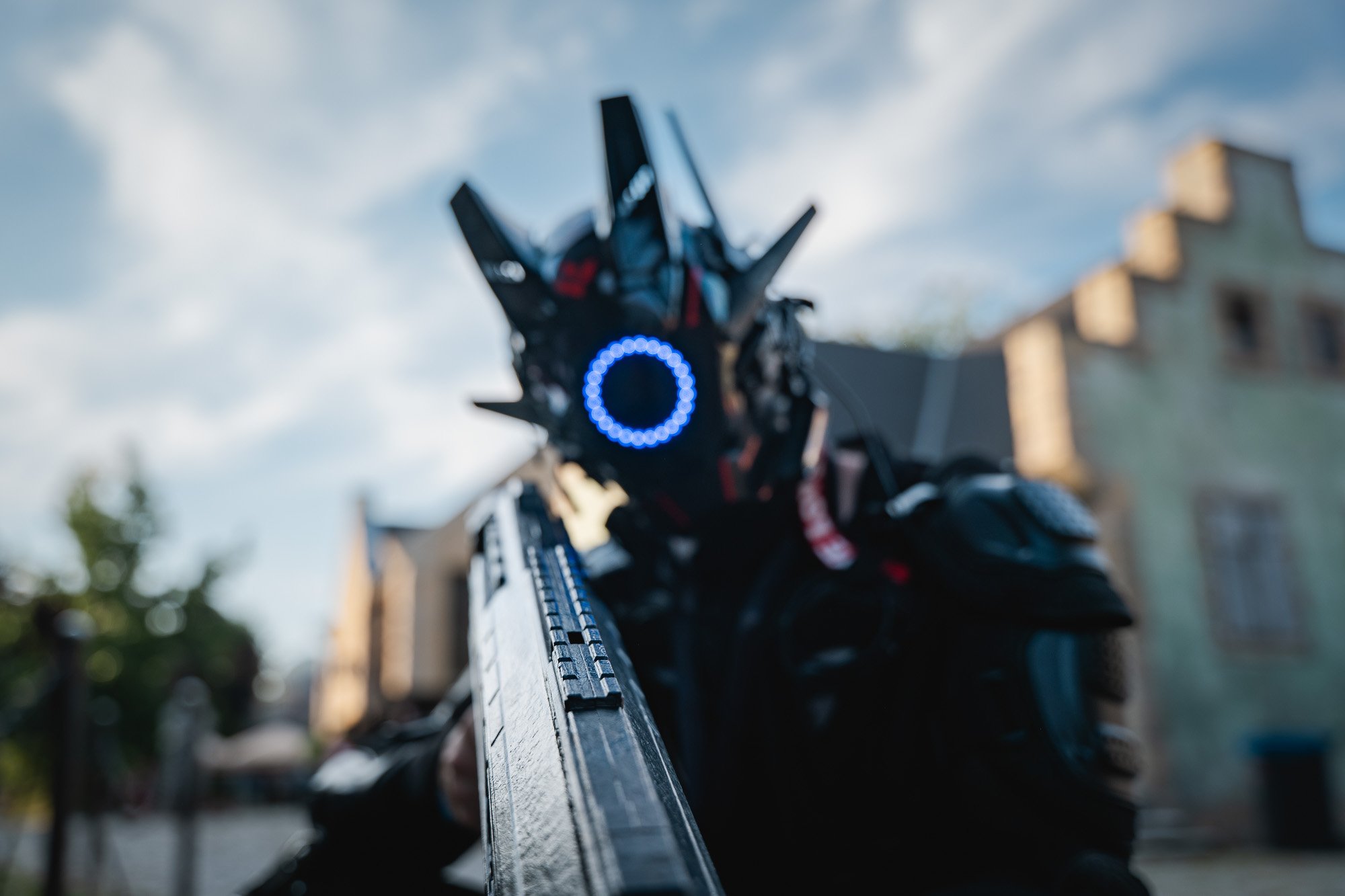 Close-up eines maskierten Charakters in schwarzer Rüstung mit spitzen Ohren, der auf eine Waffe zielt, im Hintergrund ein Gebäude und der Himmel.