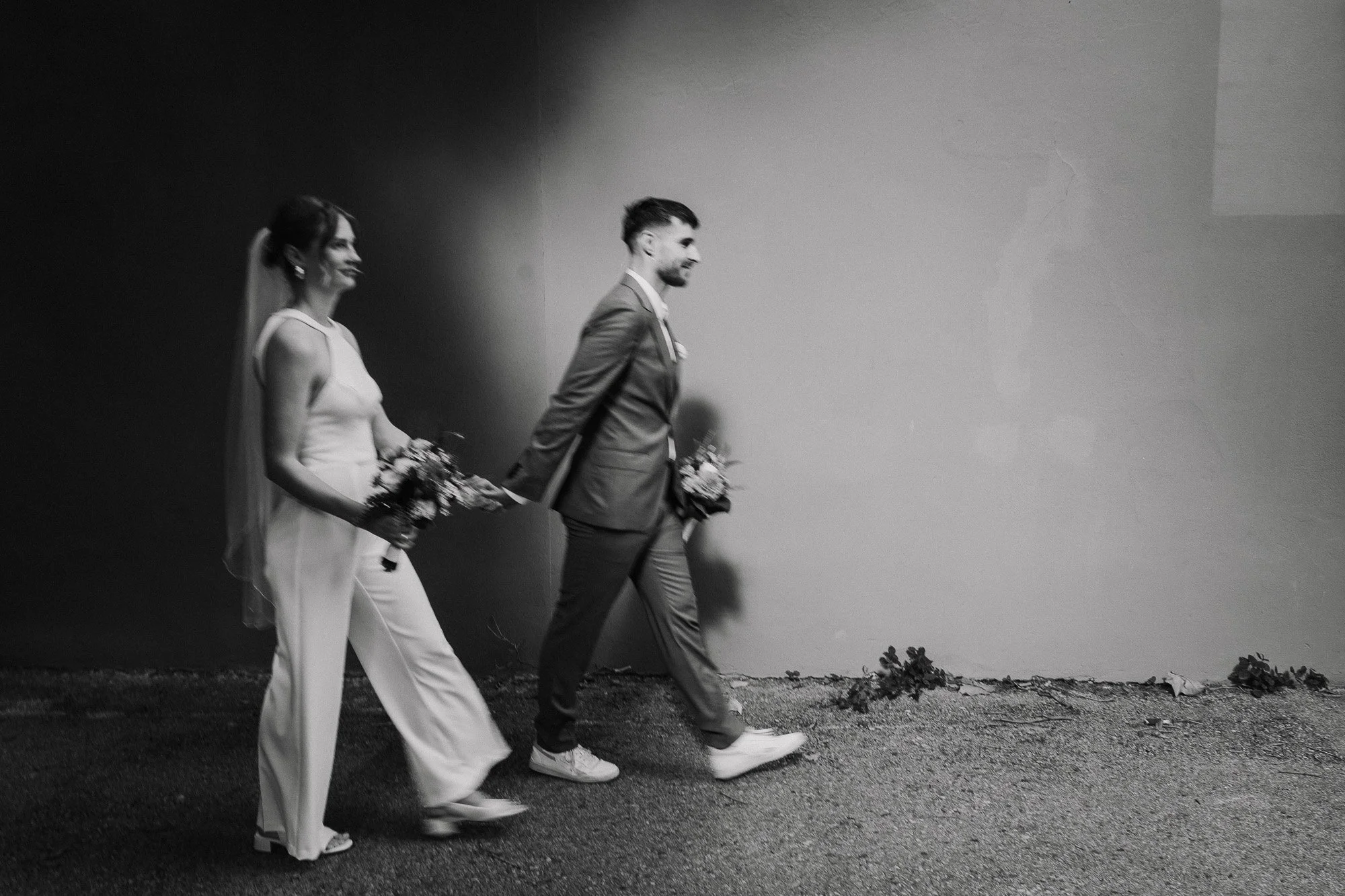 Schwarz-weiß-Foto eines verheirateten Paares, das bei ihrer Hochzeitszeremonie einander die Hände hält, während sie lächeln, mit Blumen in den Händen, vor einer Wand, während sie ihren Schritt gehen.