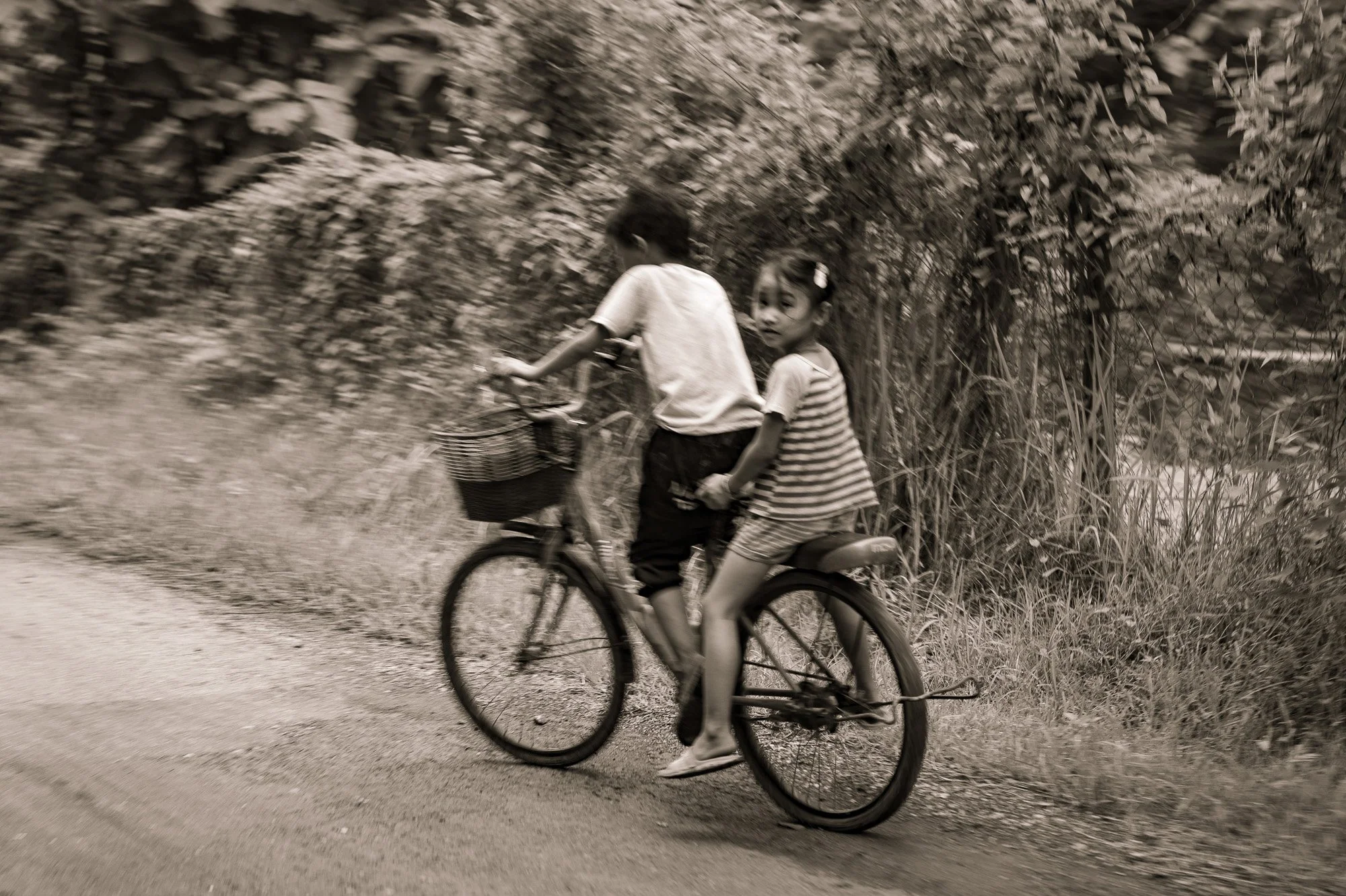 Ein Junge fährt Fahrrad, auf dem Rücken eines Mädchens, auf einem schmalen Weg, umgeben von Büschen und Bäumen, in einer ländlichen Umgebung.