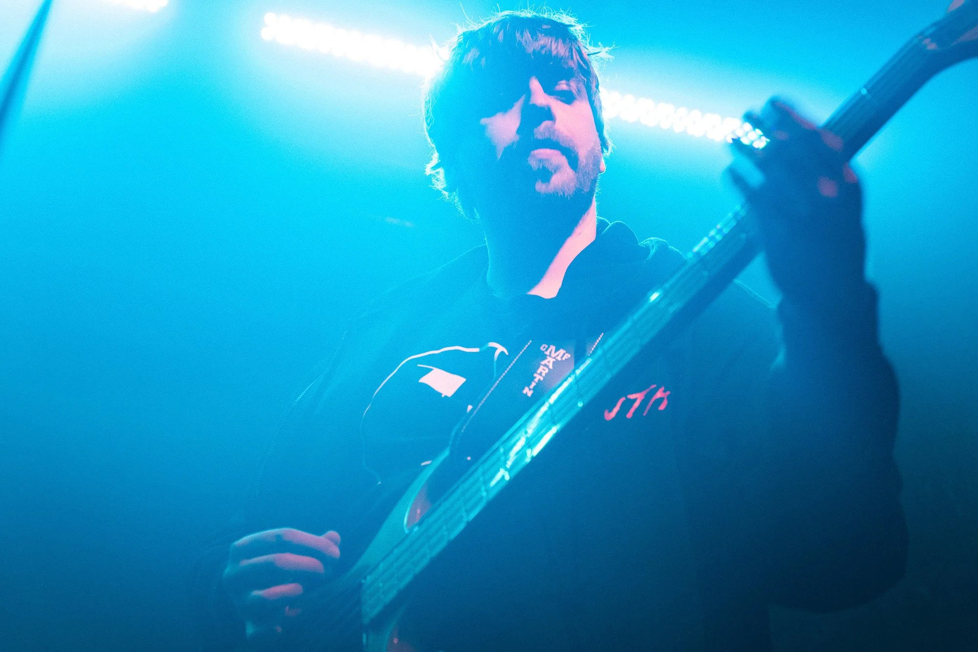 Ein Mann spielt Gitarre auf einer Bühne, umgeben von blauen Lichtern. Er hat braunes, ungepflegtes Haar und trägt dunkle Kleidung.