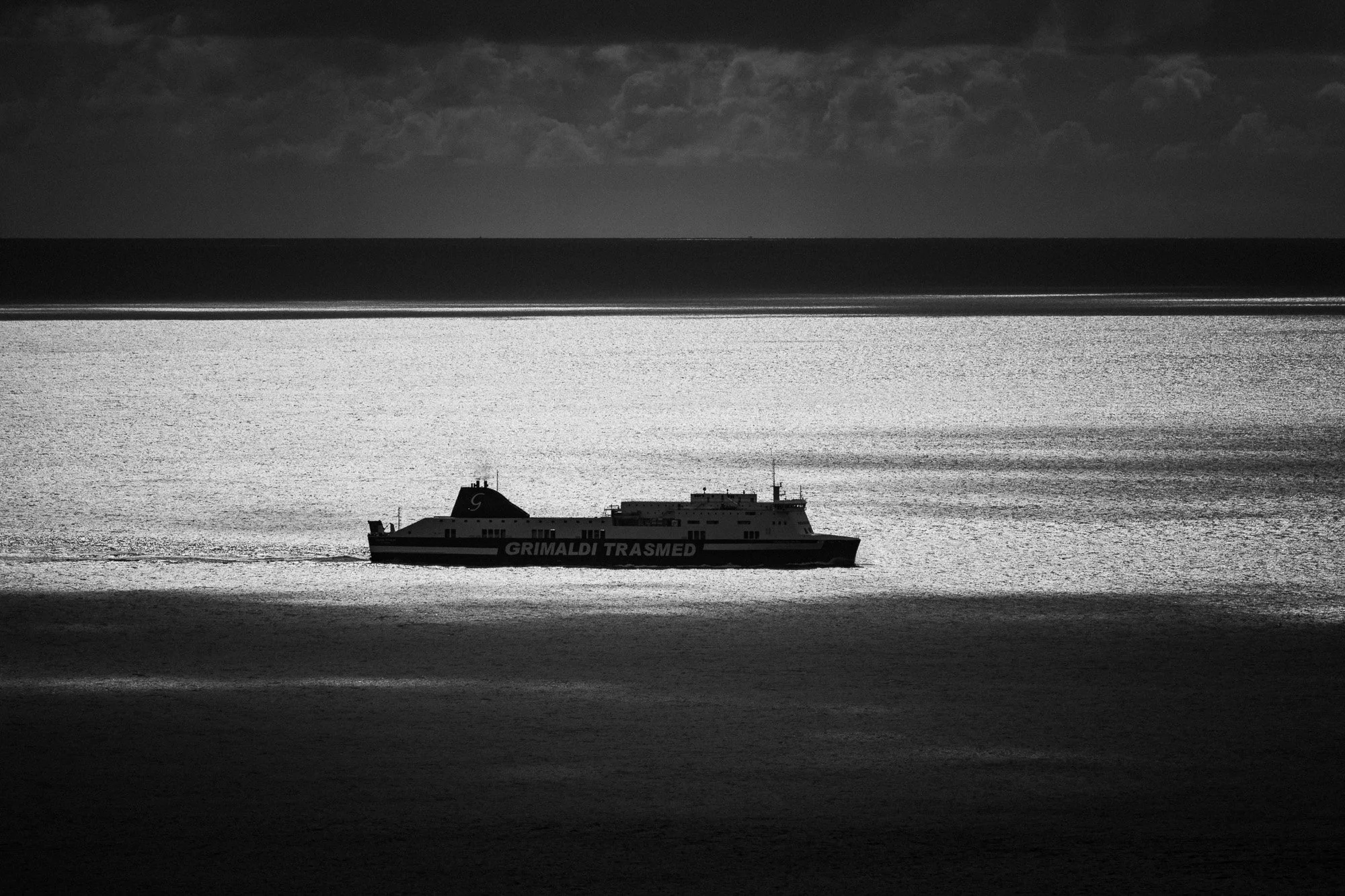 Schwarz-weiß-Foto eines Passagierschiffs mit der Aufschrift "Grimaldi Trasmed" auf ruhigem Wasser, dunkler Himmel im Hintergrund.