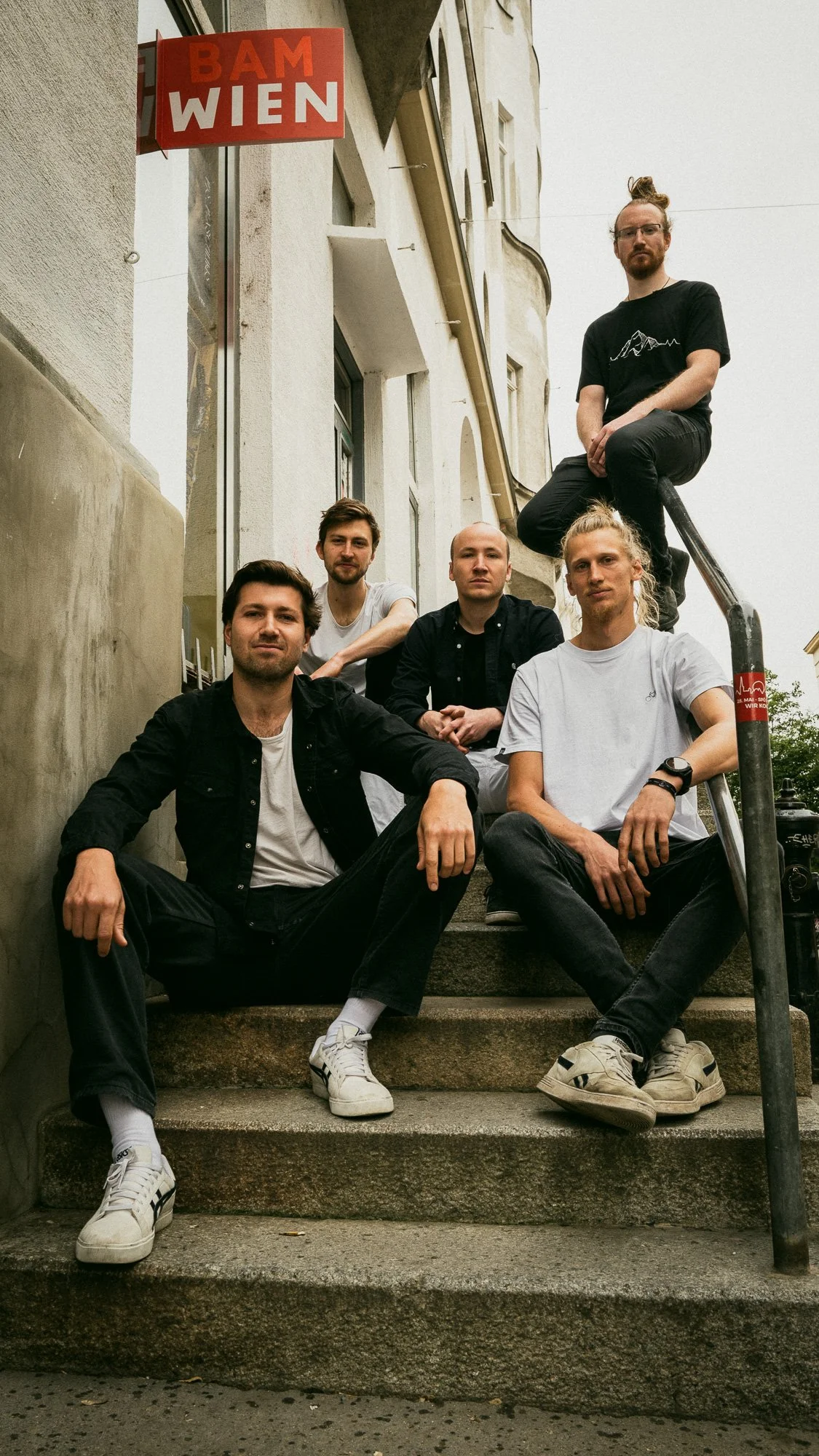 Fünf junge Männer sitzen und stehen auf den Steintreppen vor einem Gebäude, mit einem roten und weißen Schild 'BAM WIEN' oben links im Bild.